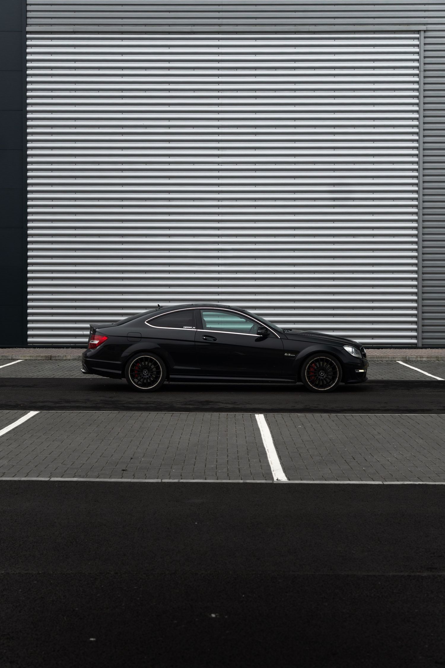 Black Mercedes-Benz C63 AMG coupe with black alloys parked against corrugated metal wall.
