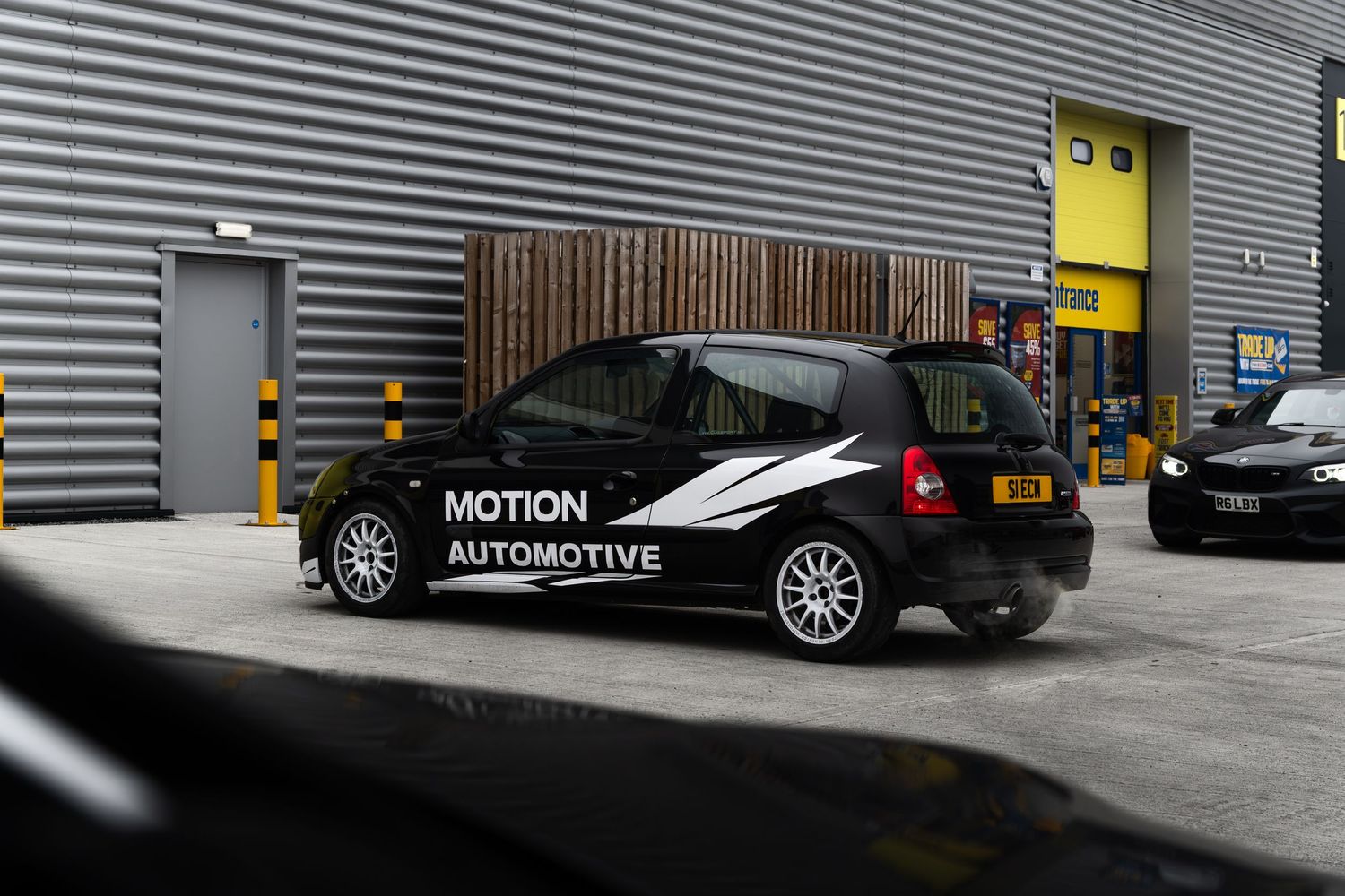 Motion Automotive branded black Renault Clio photographed from the rear three-quarter angle at an industrial unit.