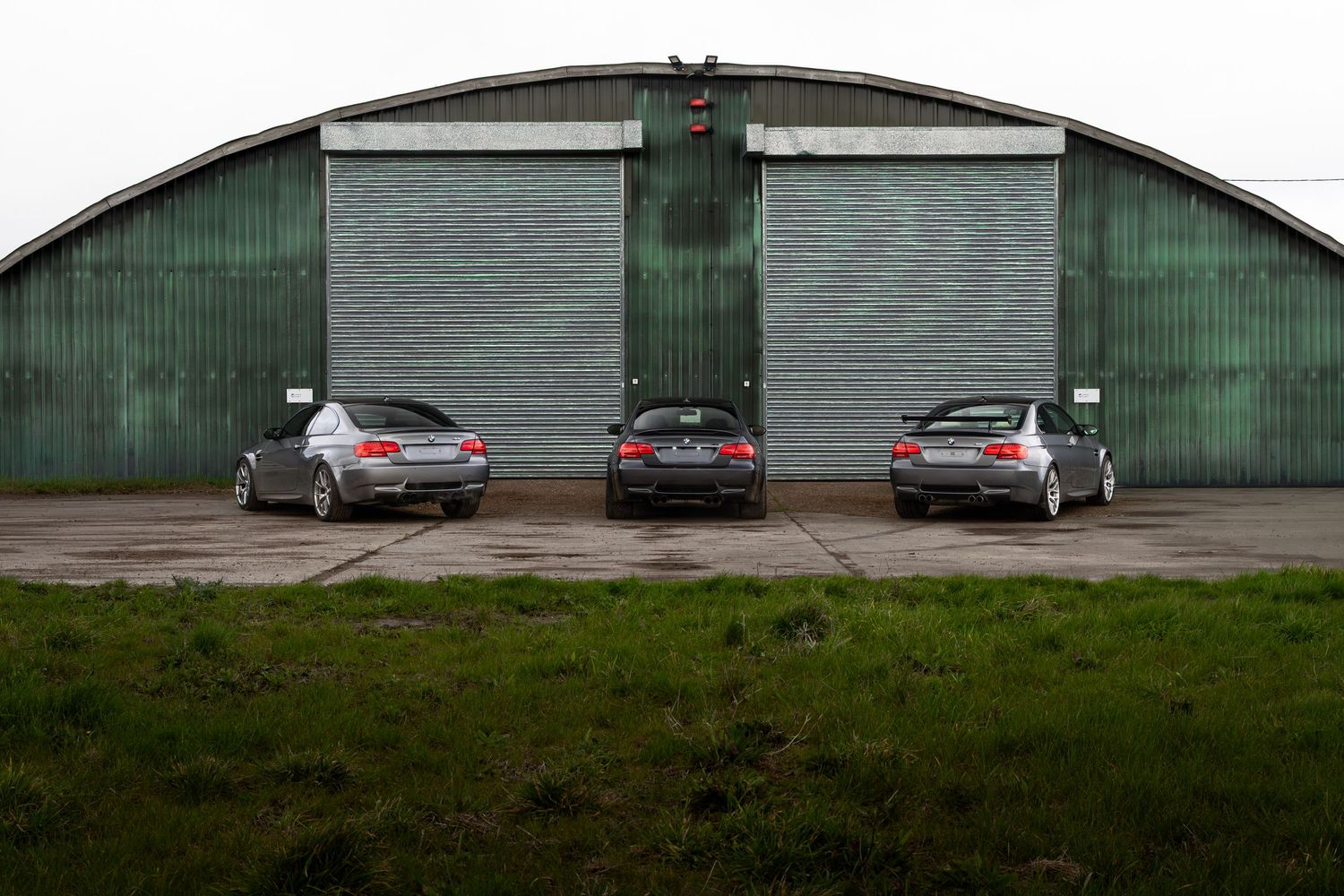 Rear view of three BMW M3 coupes lined up before green industrial hangar doors.