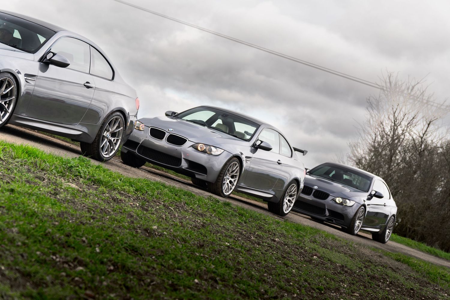 Three silver BMW M3 coupes posed on a grassy hill showcasing different generations under a cloudy sky.