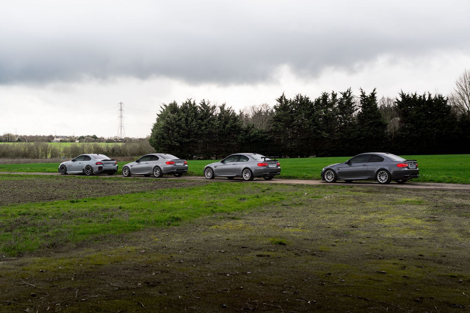 Four silver and gray BMW M3 coupes lined up on a grassy field under an overcast sky.