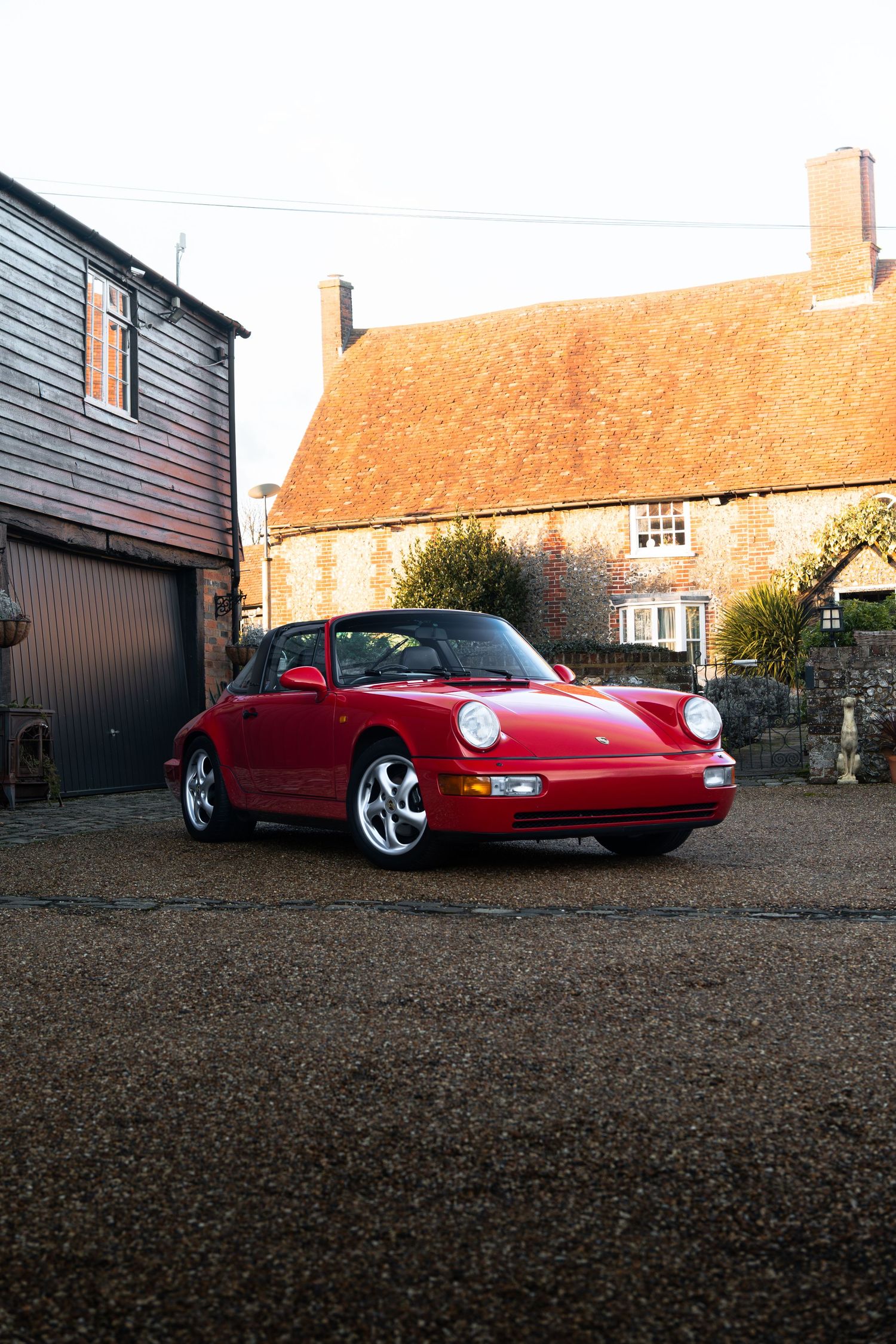 Classic red Porsche 911 Carrera photographed in front of a traditional English brick farmhouse at golden hour.