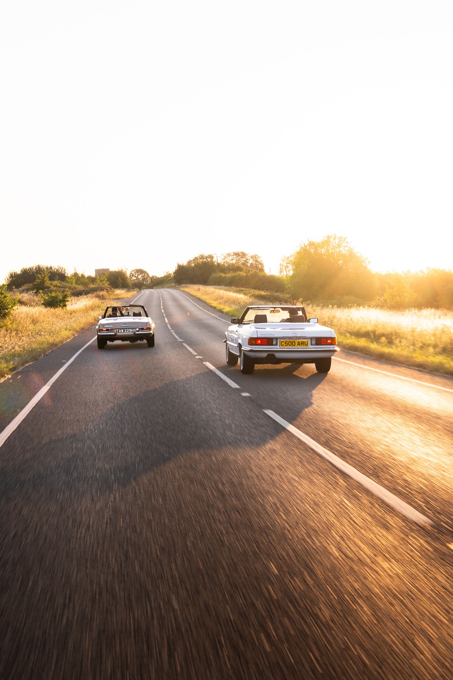 Two classic convertible cars driving on a scenic road at golden sunset hour.
