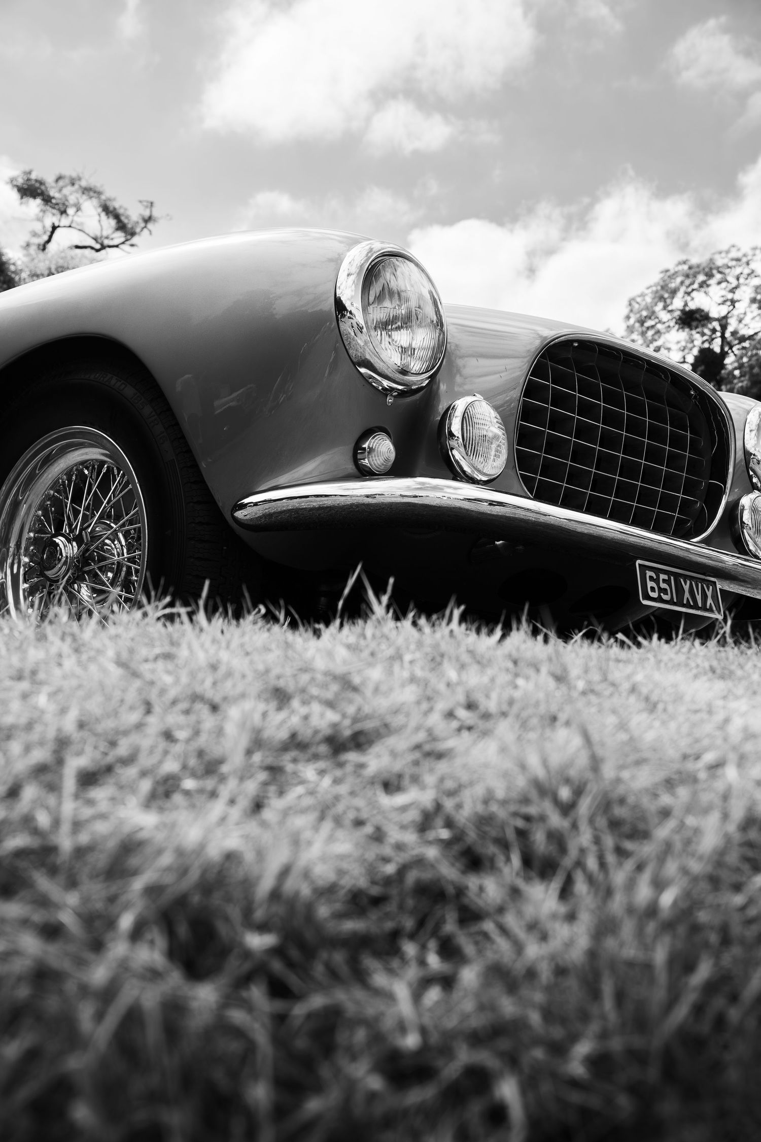 Black and white low-angle photo of a classic vintage sports car on grass, featuring chrome headlights and wire wheels.