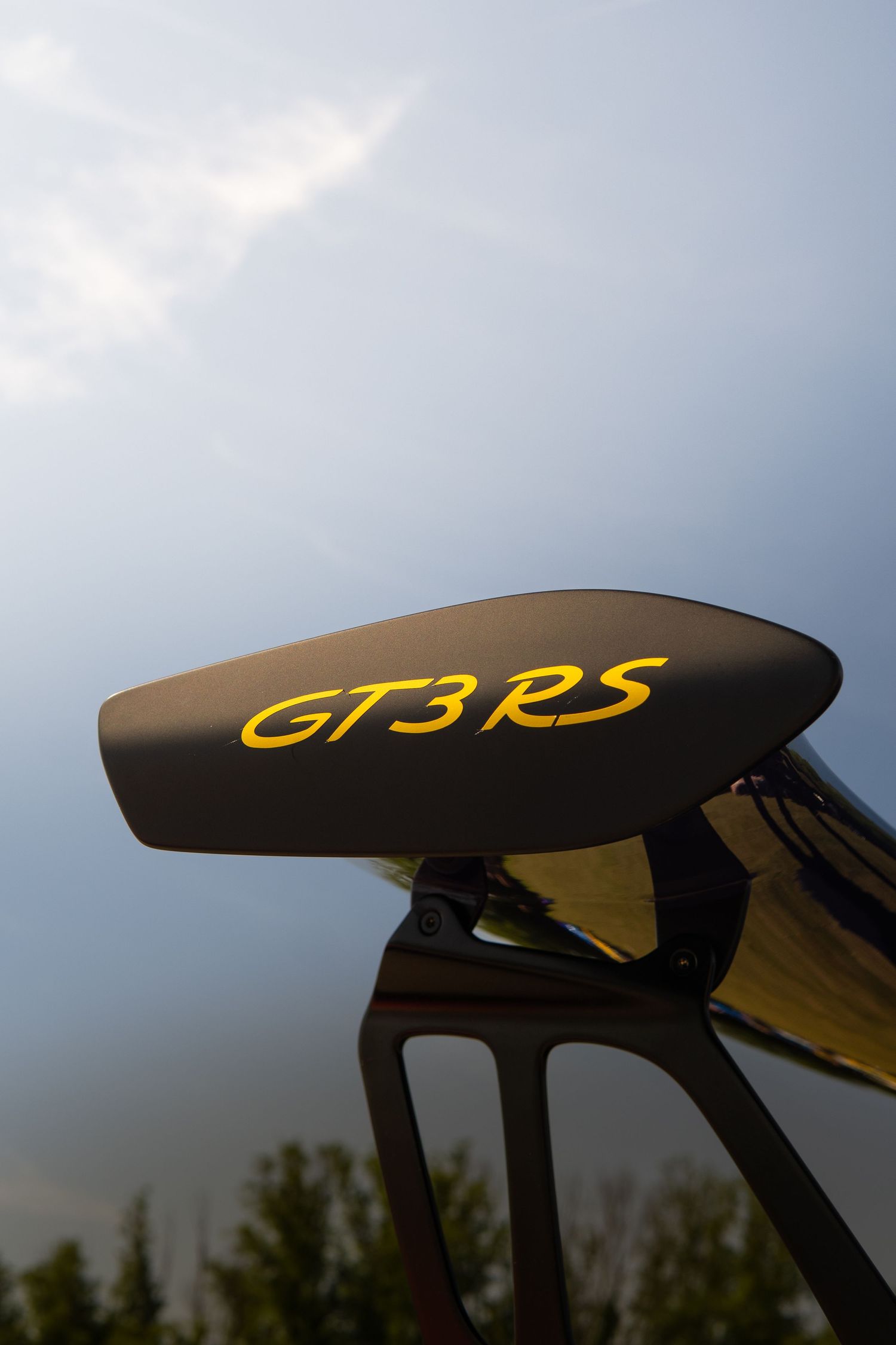 Porsche GT3 RS rear wing spoiler with yellow lettering against a dramatic cloudy sky.