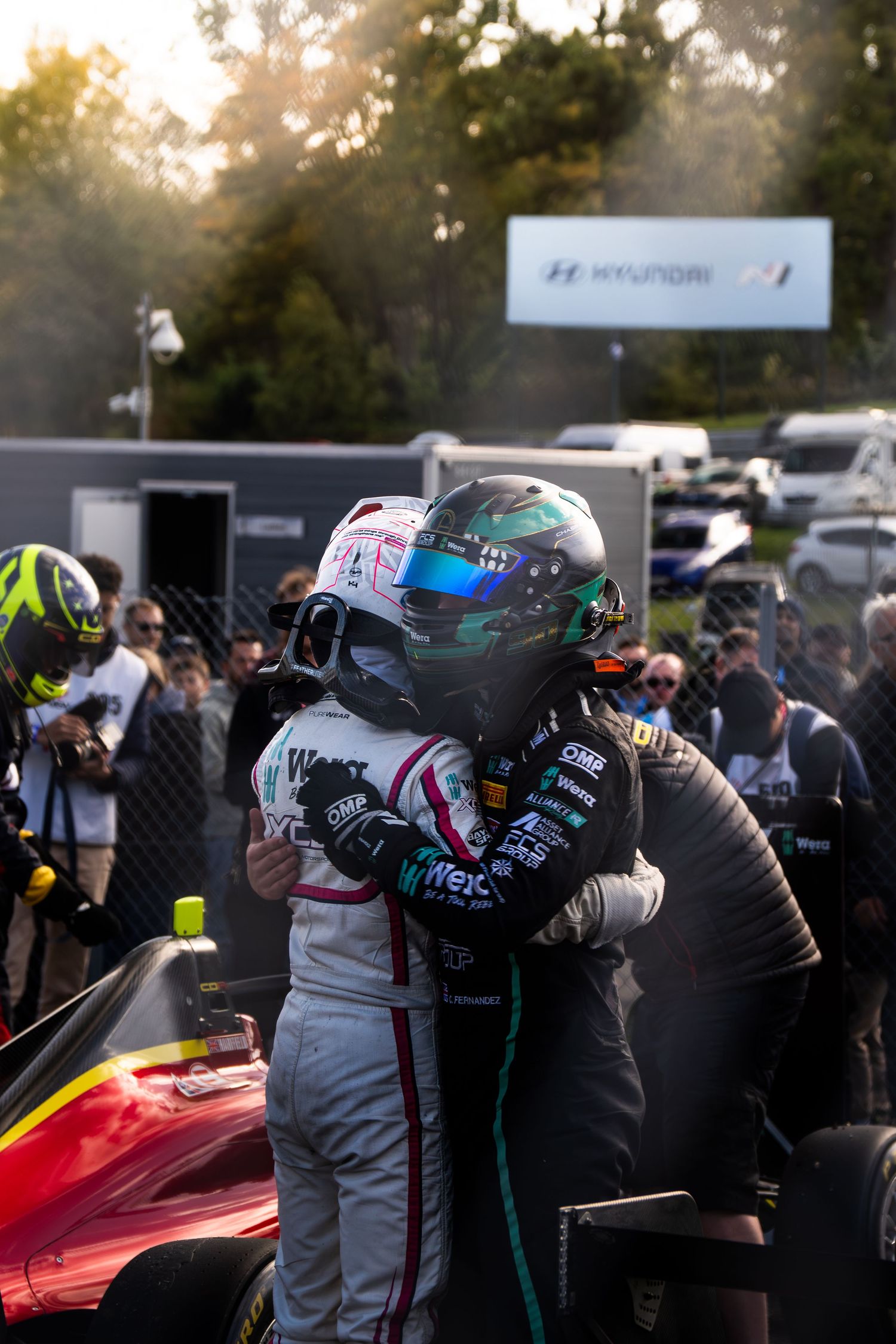 Two racing drivers in helmets embrace on the podium at a motorsport event.