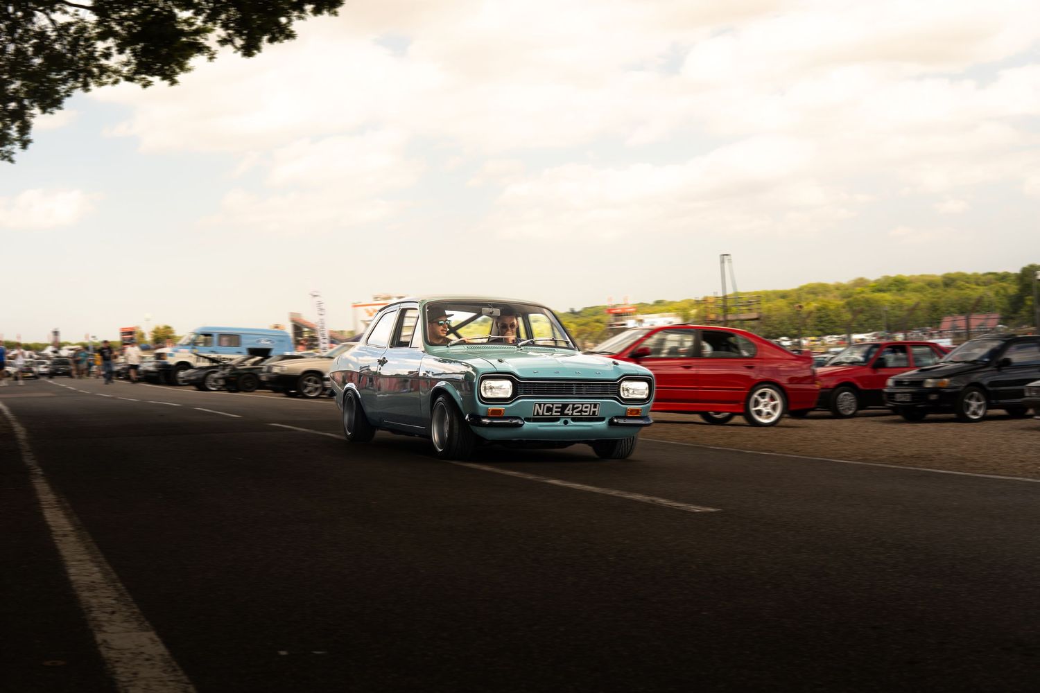 Classic light blue Mk2 Ford Escort driving through a car meet parking area at dusk.