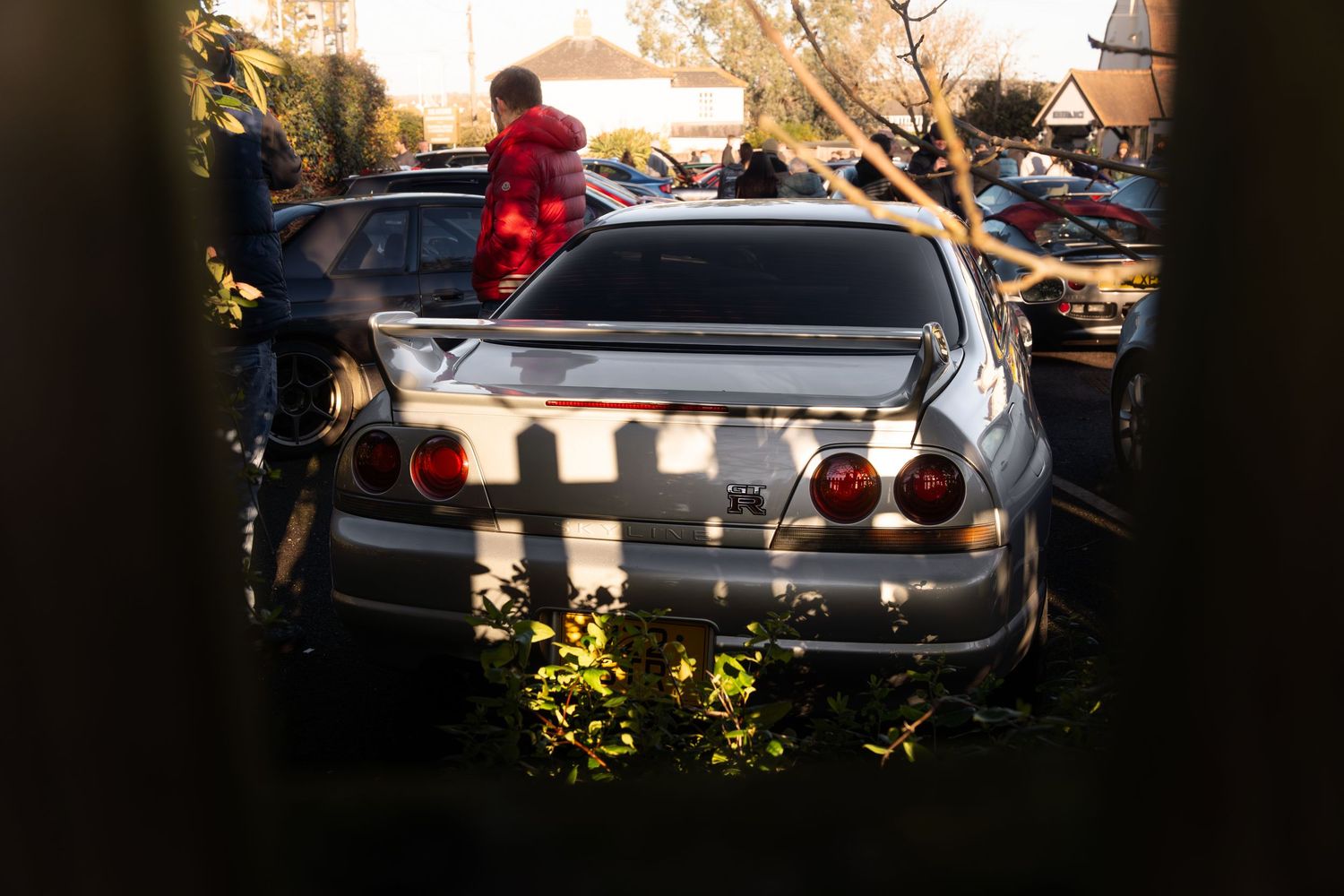 Silver Nissan Skyline R33 GT-R viewed through foliage at a sunny car meet event.