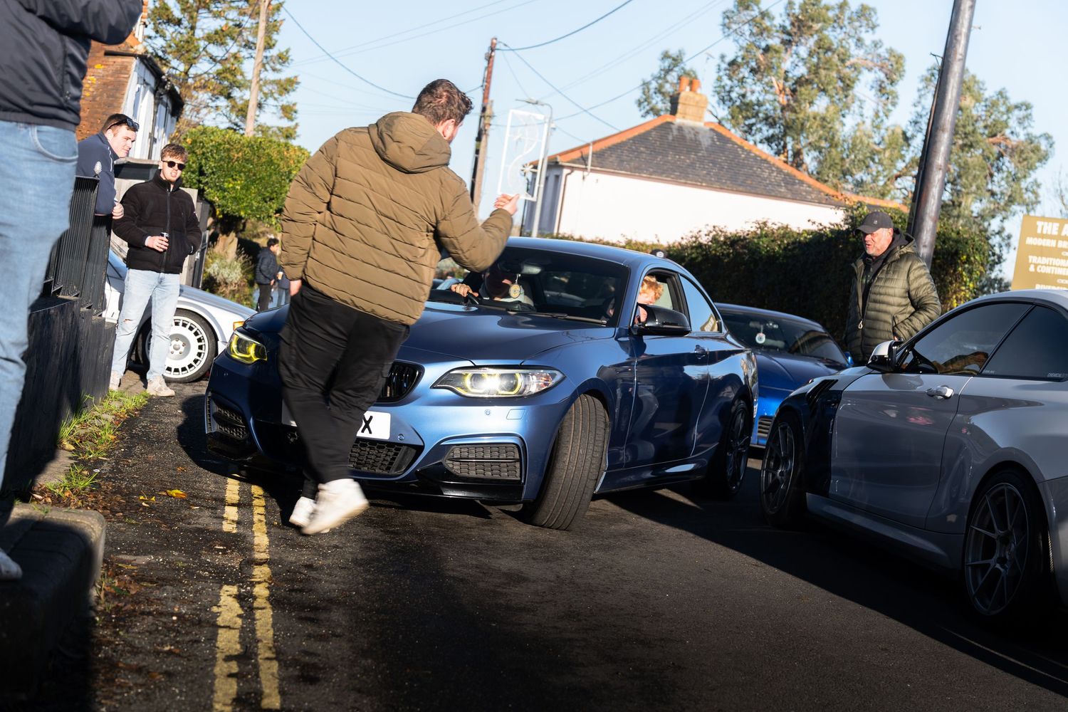 Enthusiasts gathering around a blue BMW M2 on a narrow road during an outdoor car meet event.