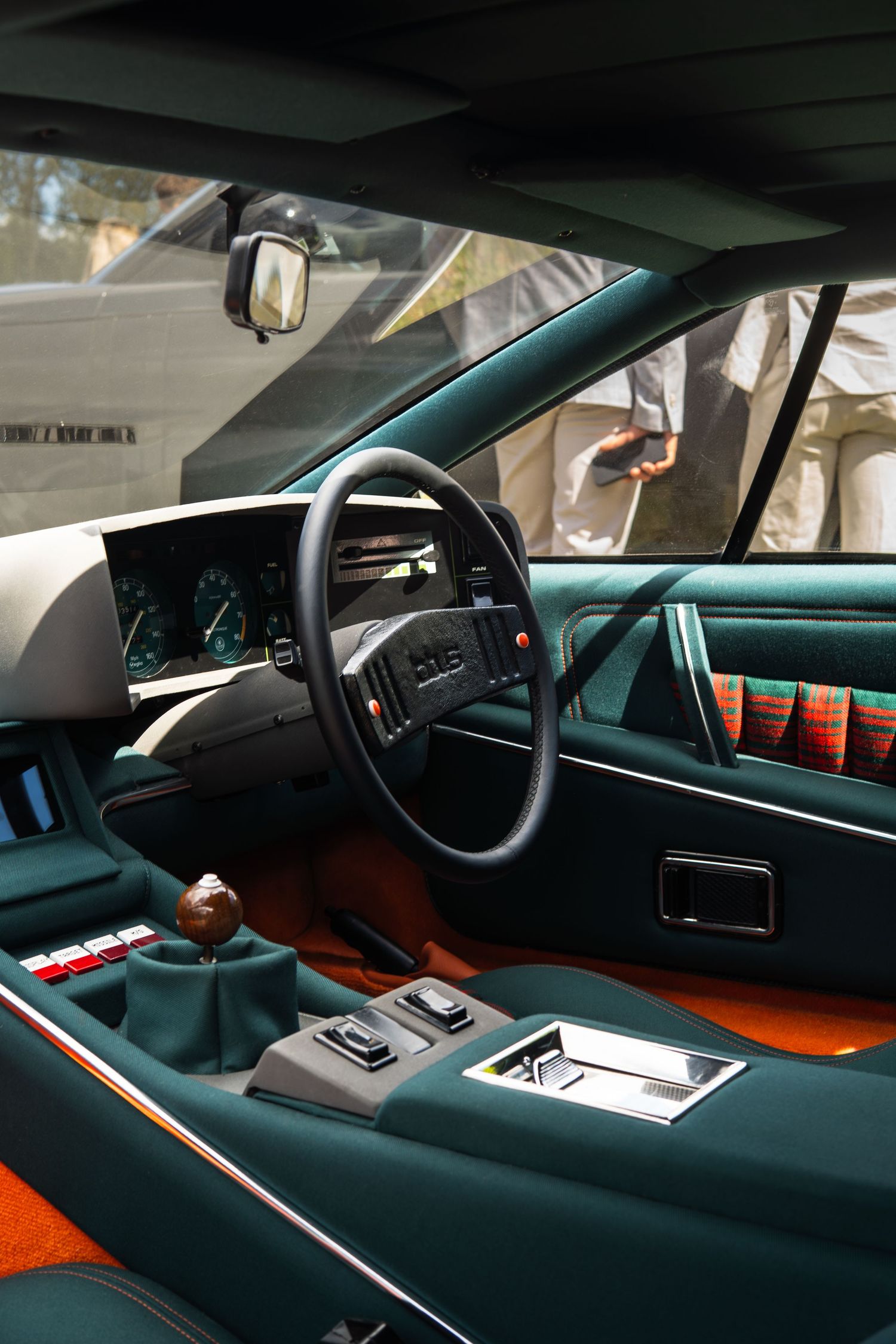 Vintage concept car interior featuring teal upholstery, plaid seats, orange carpet, and retro dashboard at a car show.
