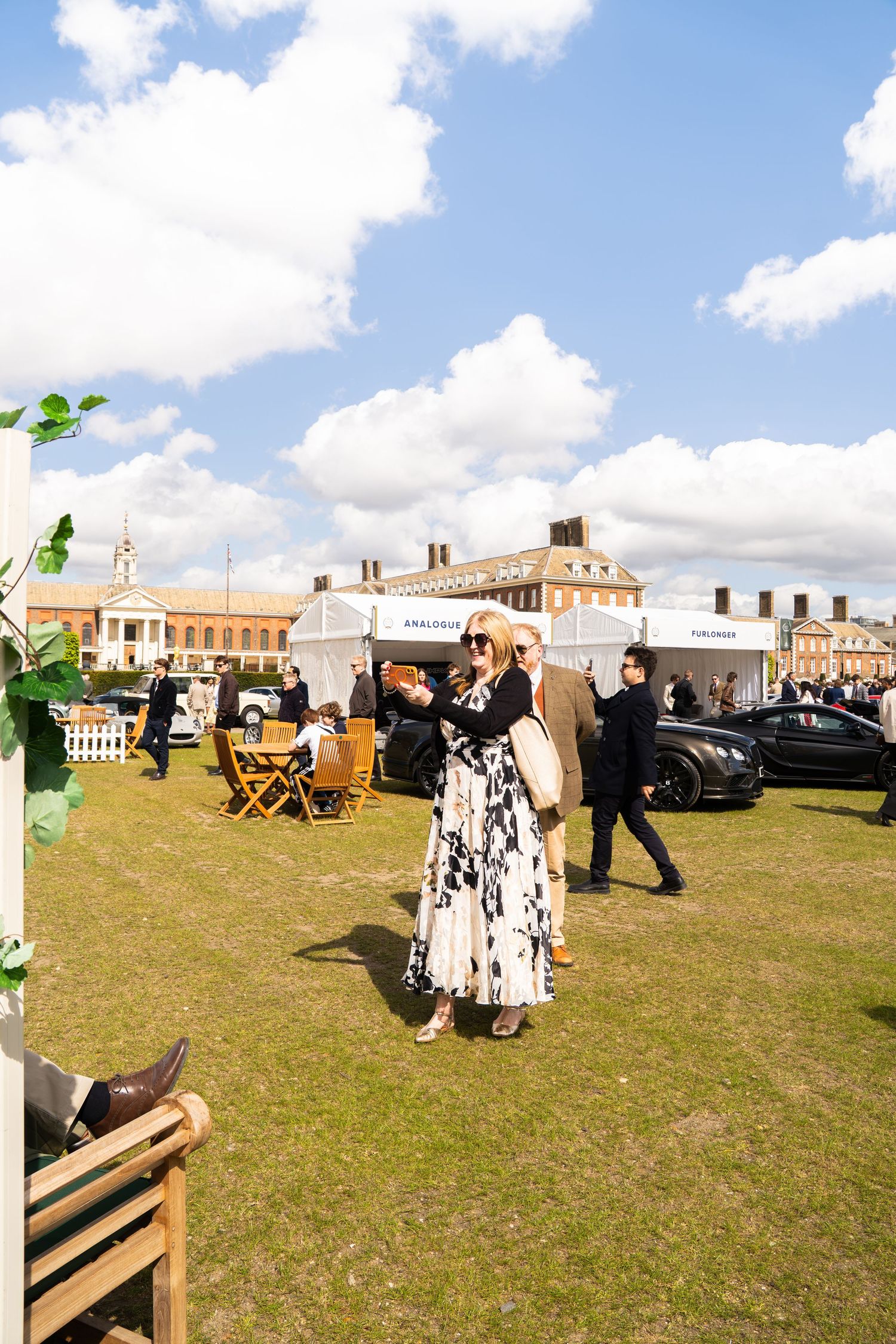 Elegantly dressed woman at an outdoor luxury event on a sunny day with historic buildings in background.