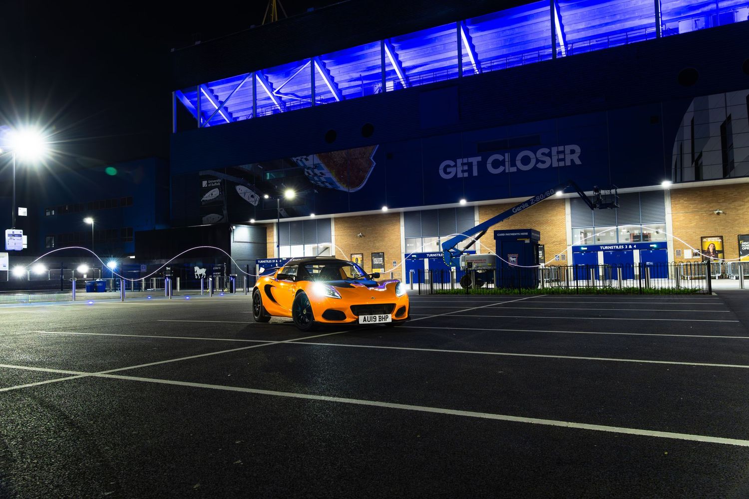 Orange Lotus Elise sports car in night-time stadium car park under blue LED lighting with GET CLOSER billboard.