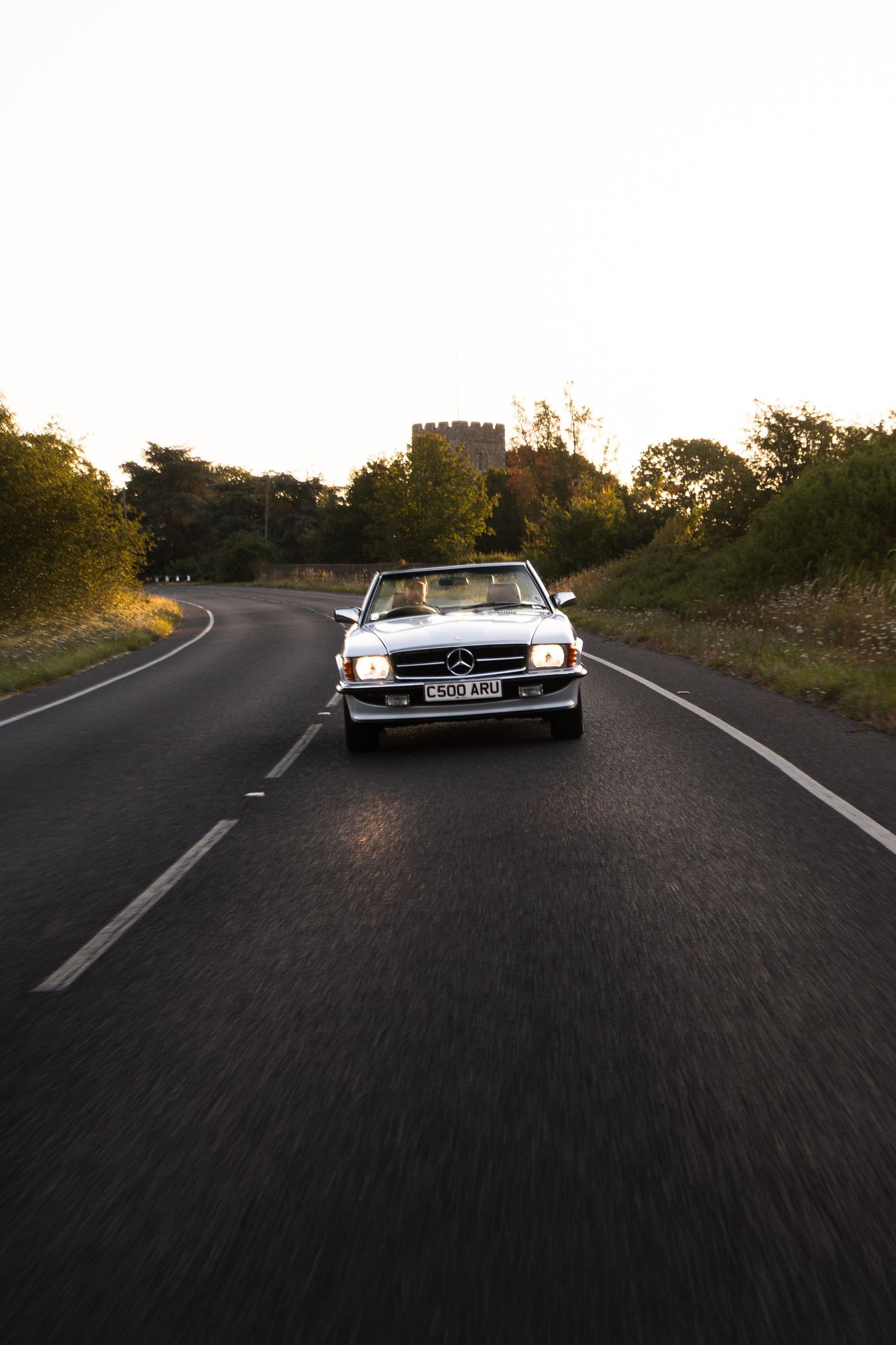 Classic Mercedes-Benz convertible driving on a winding country road at golden hour, surrounded by lush green trees.