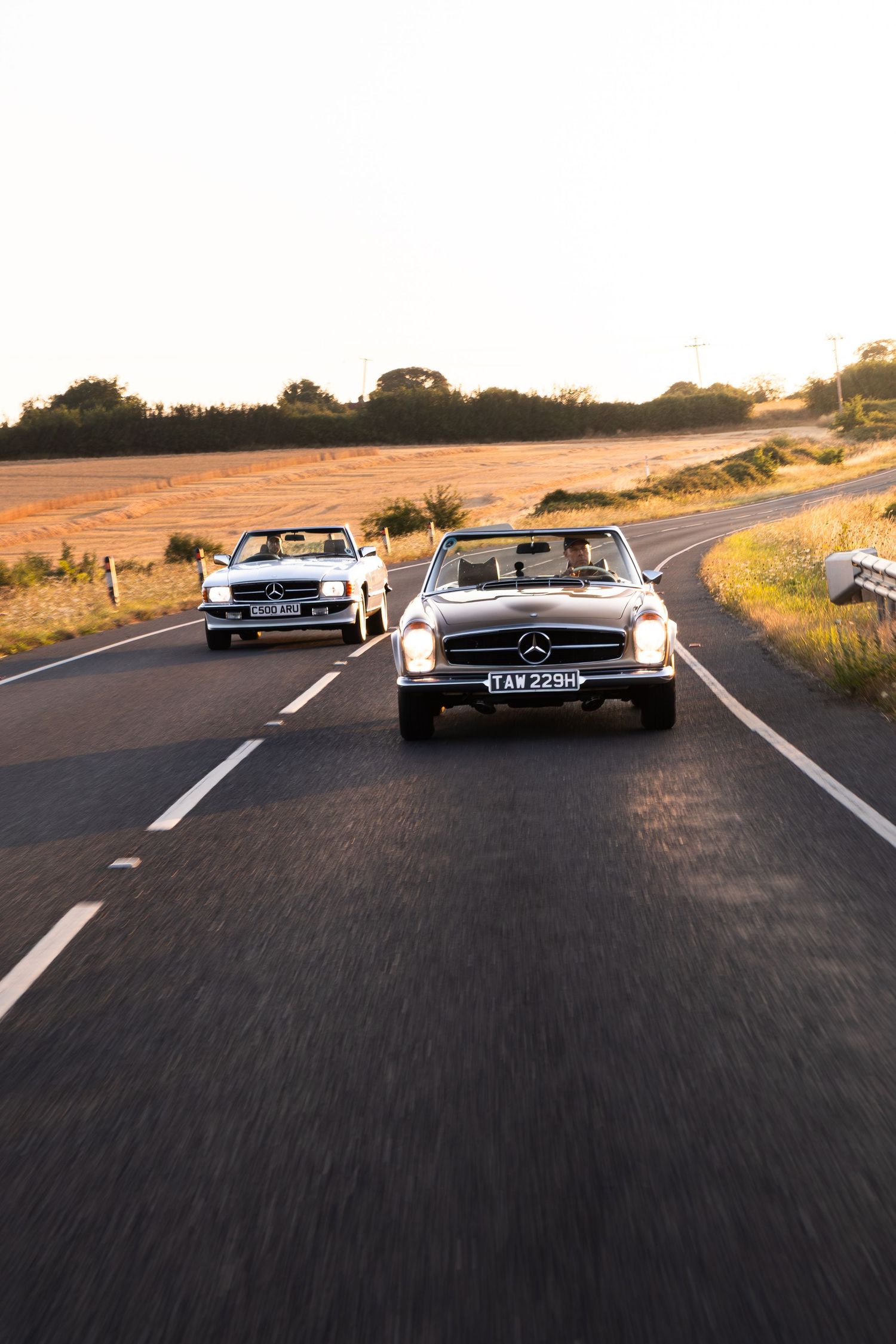 Two classic Mercedes-Benz convertibles driving on a rural road at golden hour sunset.