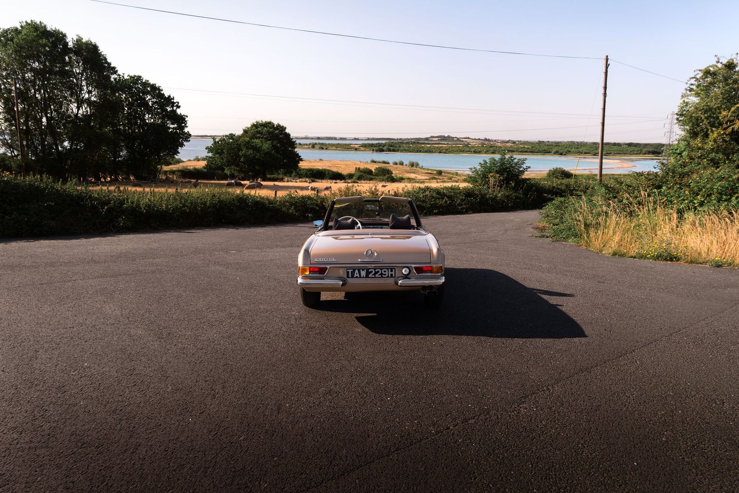 Vintage convertible car parked on a quiet road surrounded by lush greenery and a serene lake at sunset.