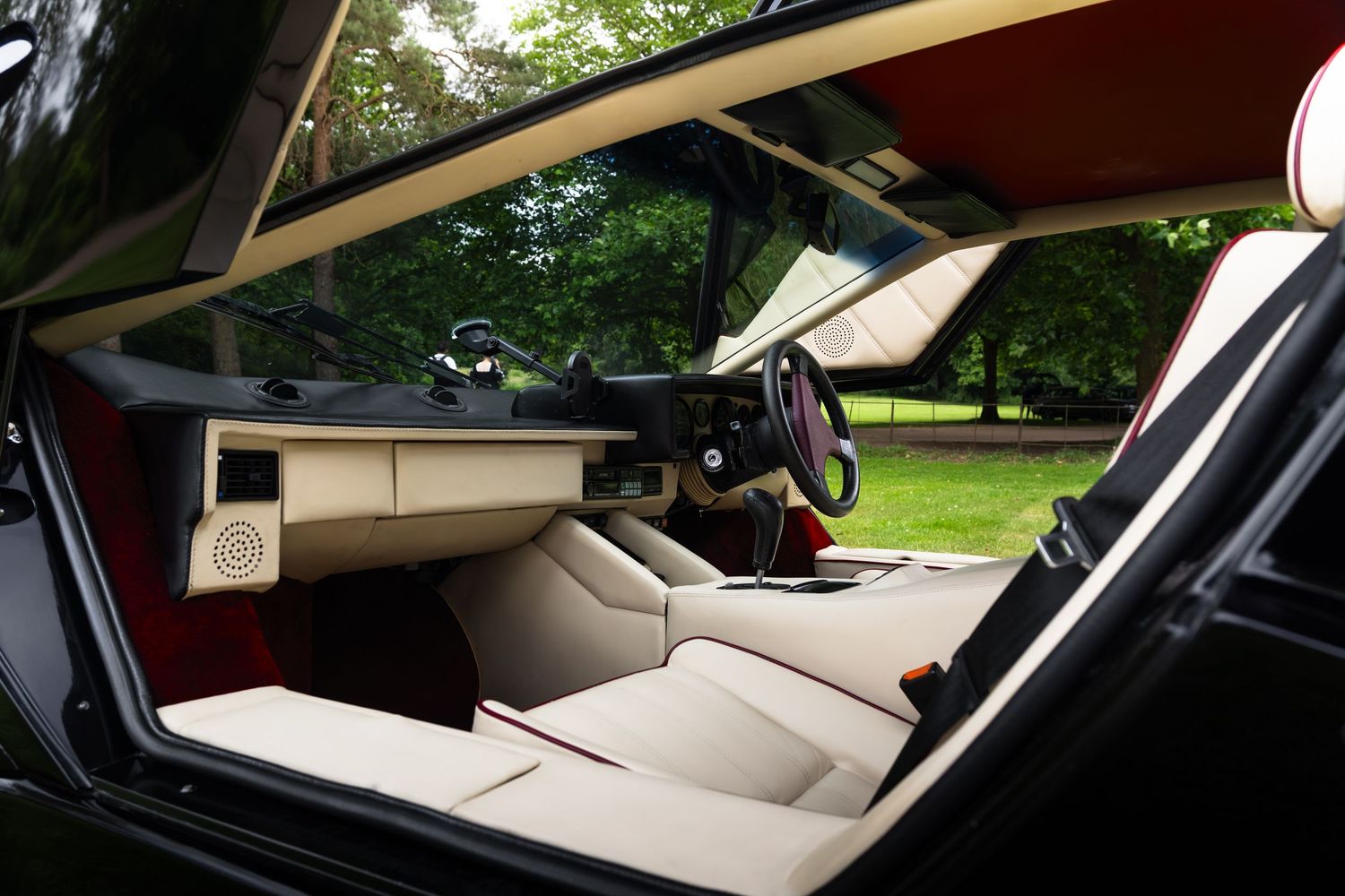 Luxury sports car interior with cream leather seats, red accents, and a sleek dashboard surrounded by lush green trees.