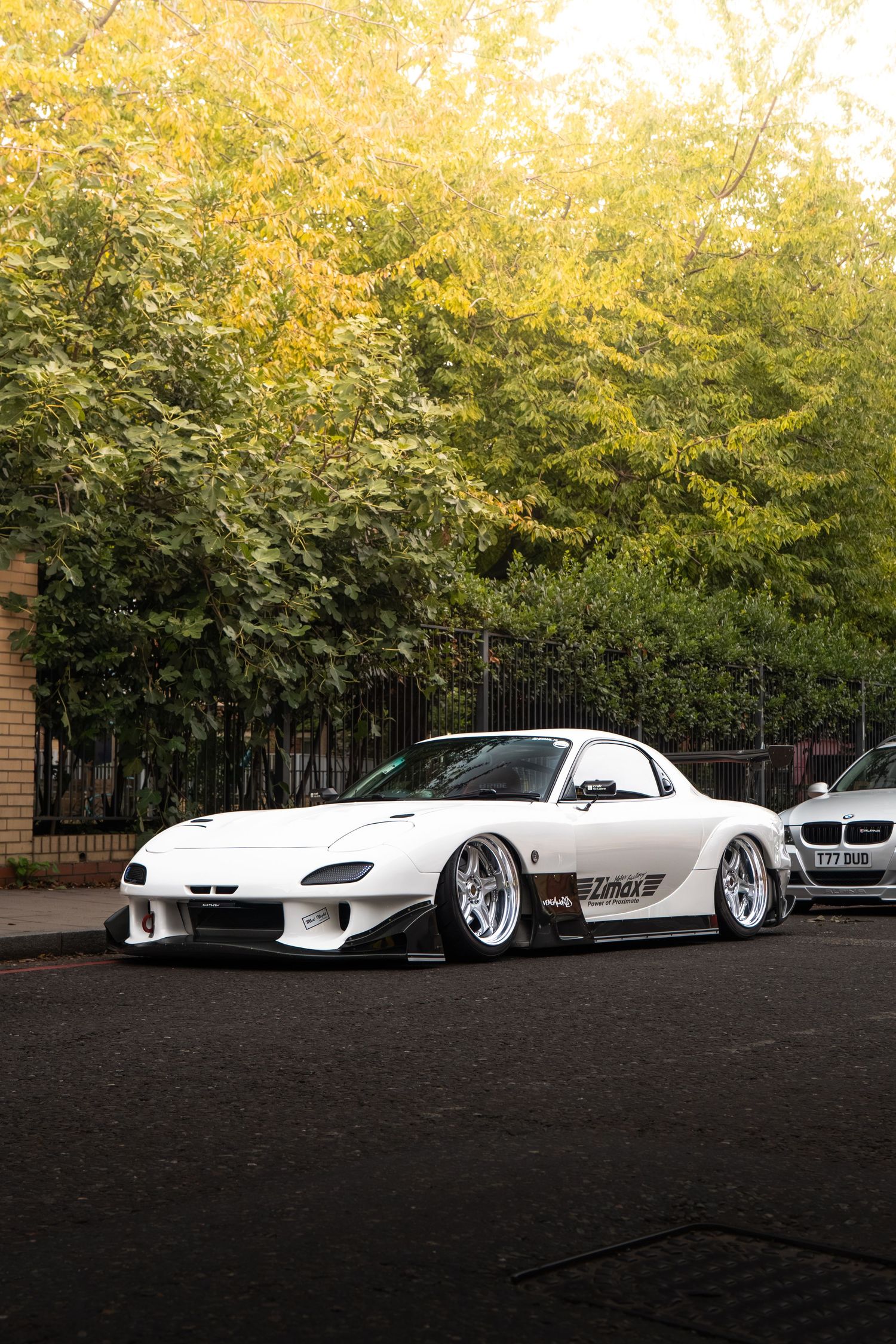 A white widebody Mazda RX-7 FD on silver wheels parked on a street lined with green trees.