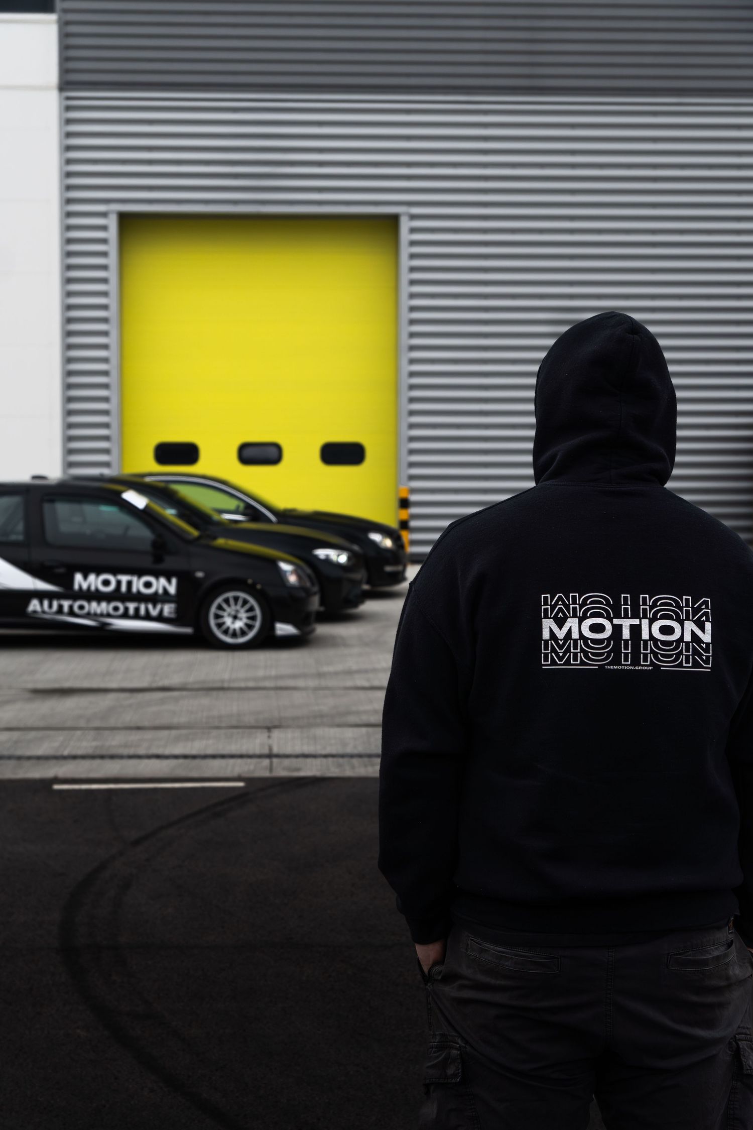 Person wearing a black Motion hoodie standing in front of Motion Automotive branded cars at a garage.
