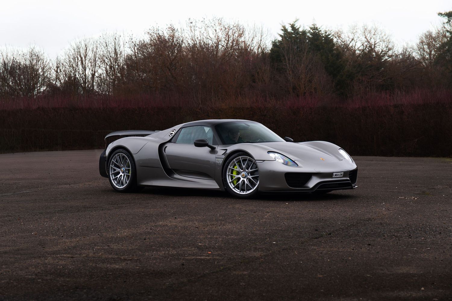 Silver Porsche 918 Spyder supercar parked on gravel with autumn trees in background.