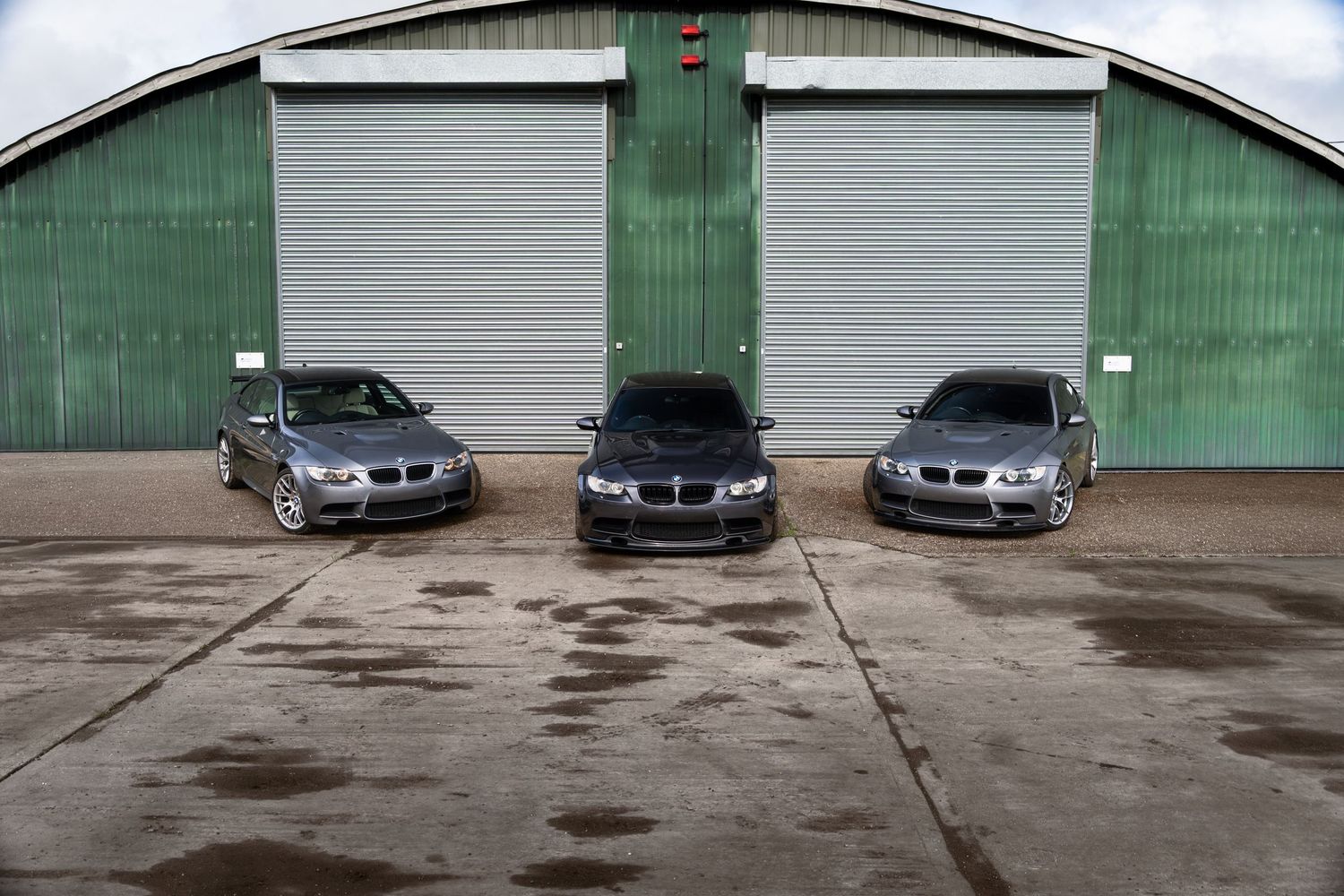 Three grey BMW M3 cars parked in front of green industrial garage doors.