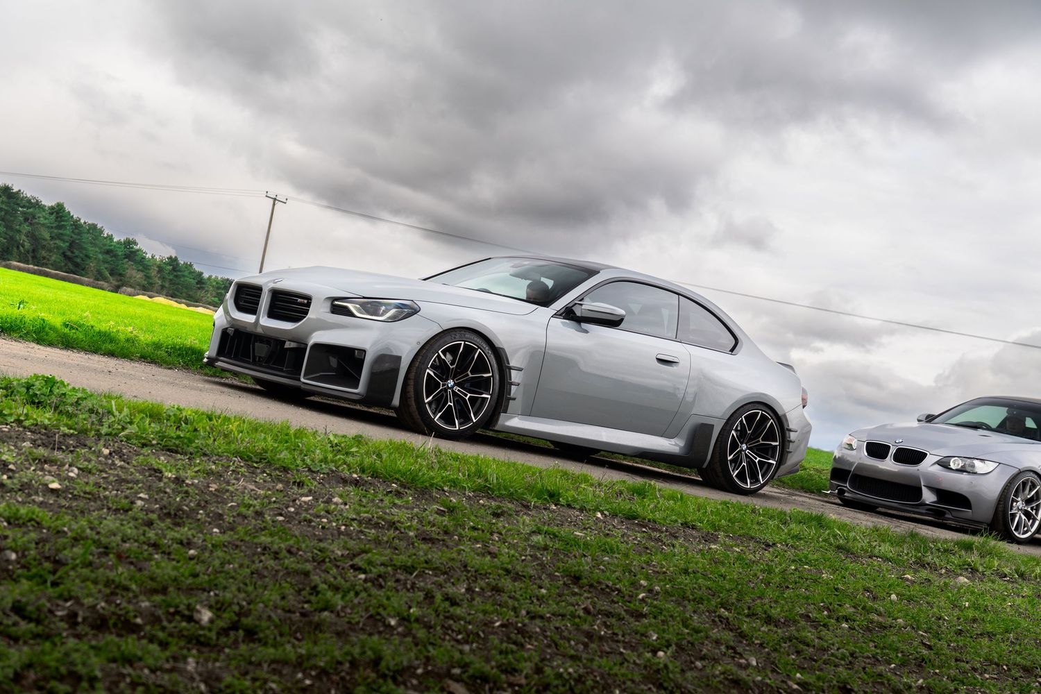 Silver BMW M2 Competition with aggressive body kit parked on a green grass roadside under overcast skies.