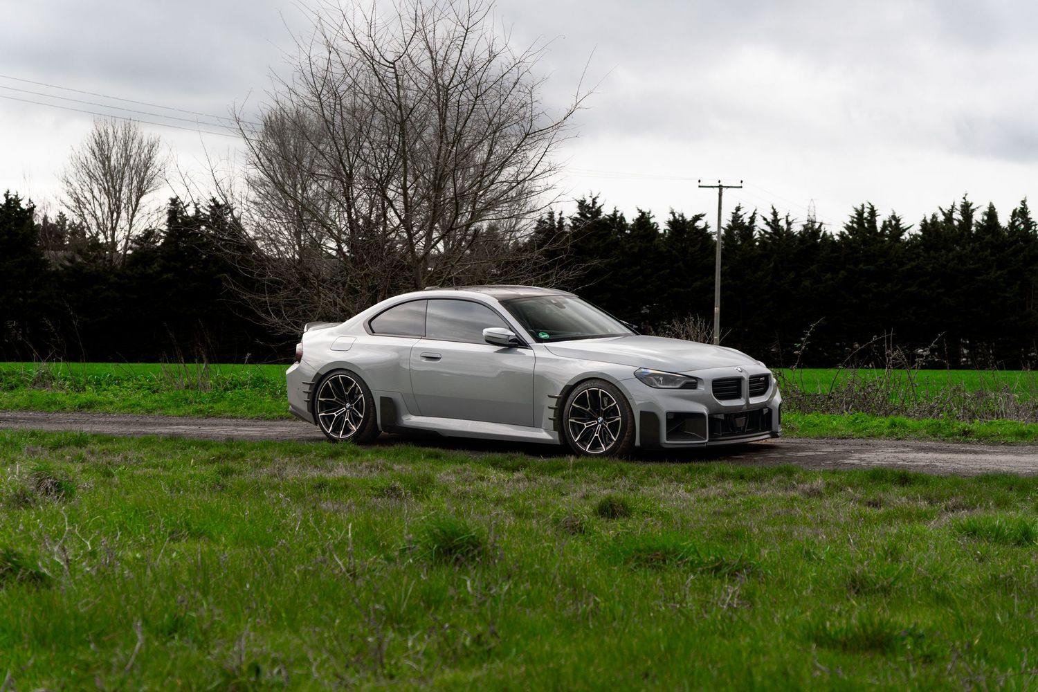 Silver BMW M2 coupe parked on a country road surrounded by green fields and trees.