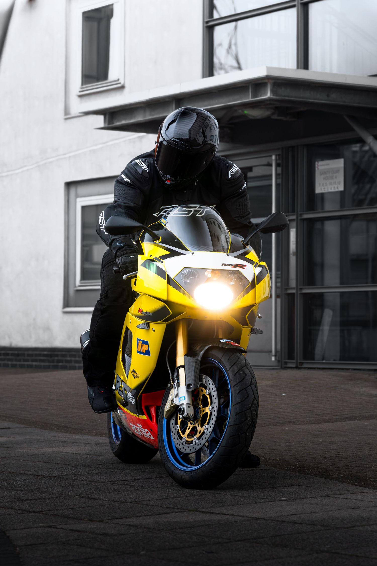 Front view of yellow Aprilia RSV superbike with glowing headlight outside building.
