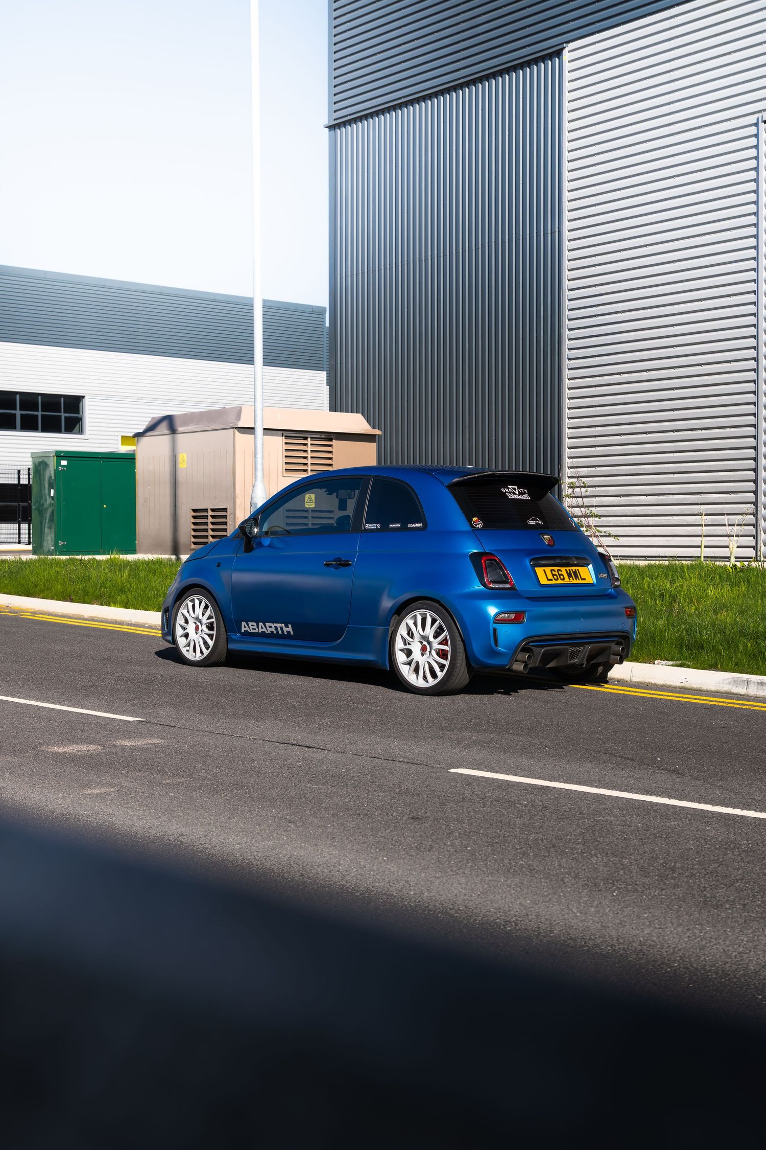 Rear three-quarter view of a blue Abarth 500 with UK number plate parked near an industrial building.