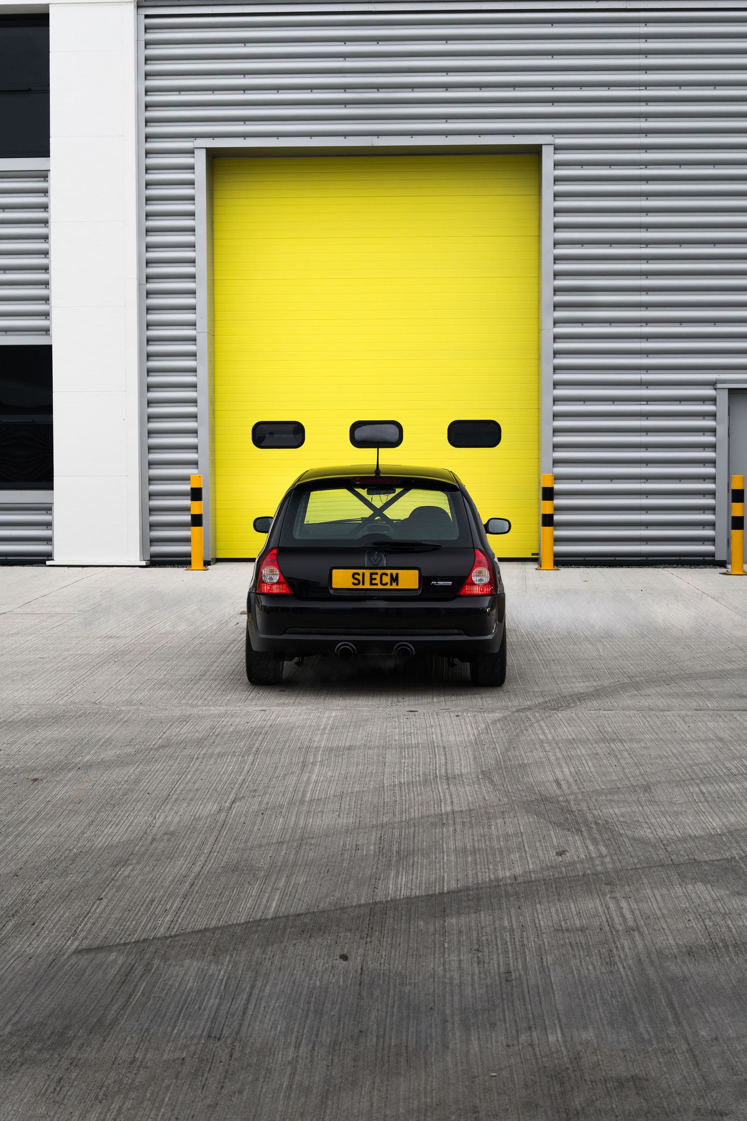 Rear view of a black Renault Clio with S1ECM plate parked in front of a bright yellow roller shutter door.