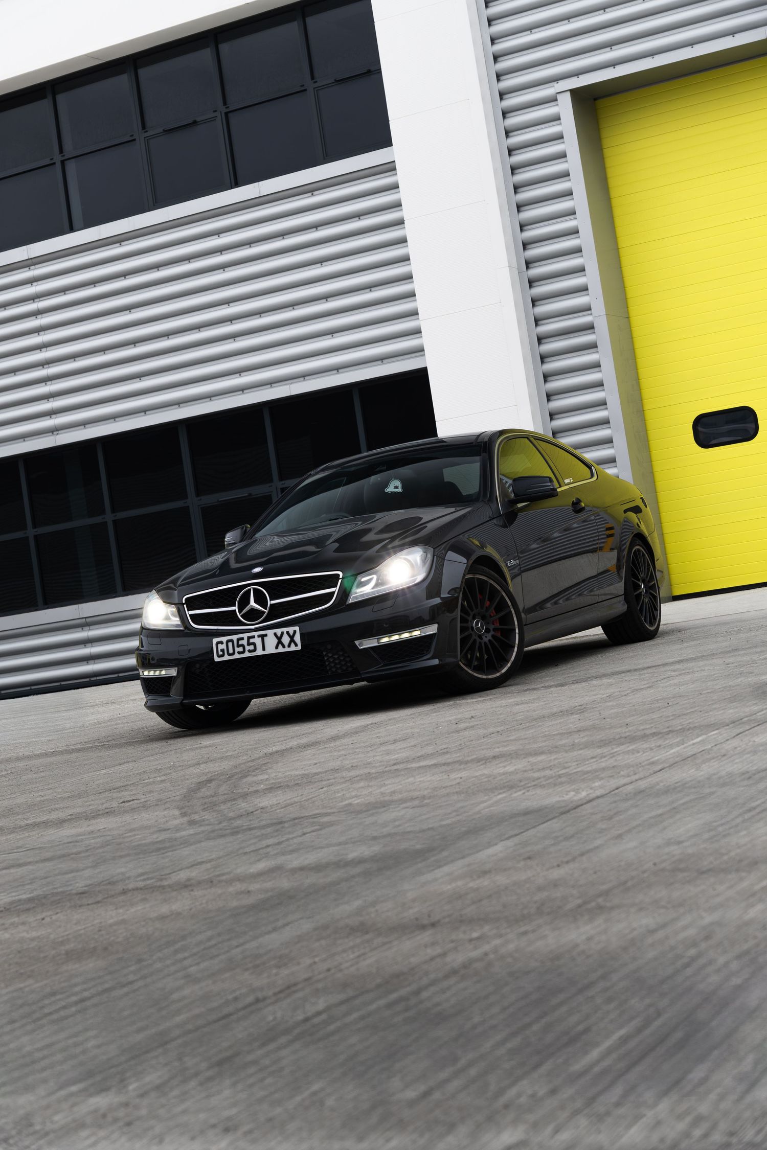 Black Mercedes-AMG C63 coupe with headlights on driving near yellow industrial garage door.