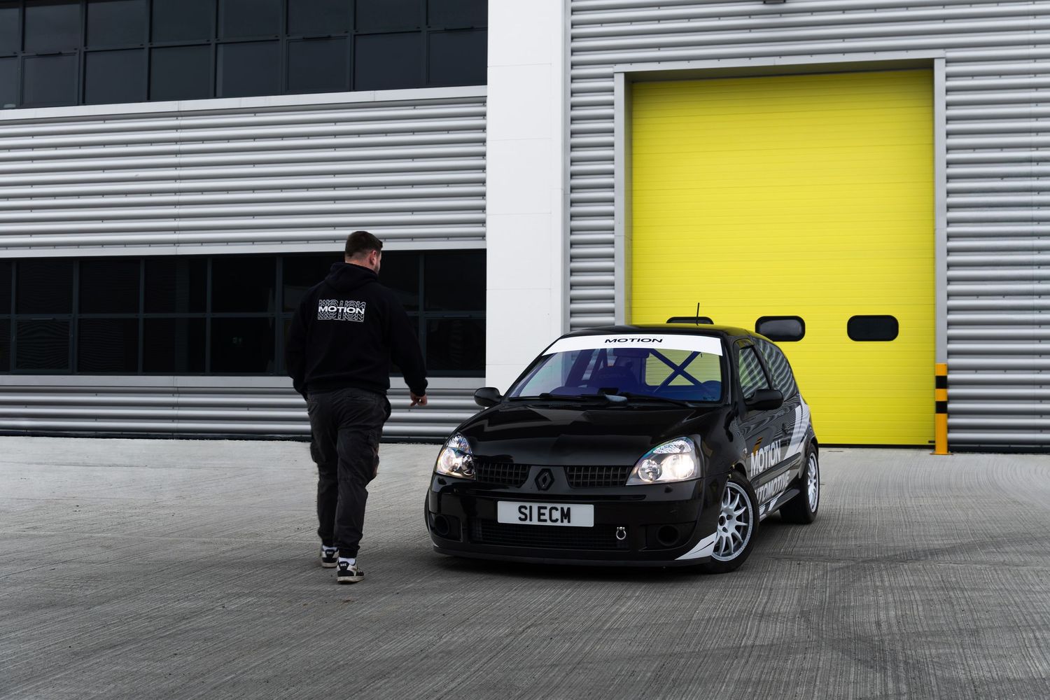 Person in black jacket walking towards a black Motion Automotive Renault Clio parked outside yellow garage doors.