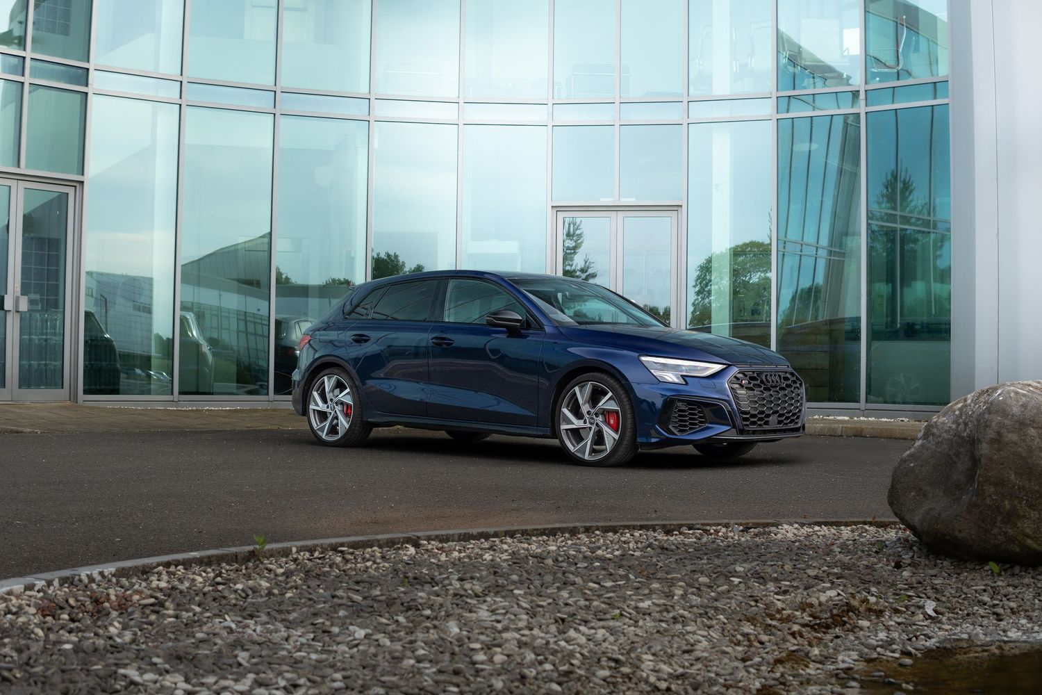 Navy blue Audi S3 sedan parked on a curved driveway beside a modern glass building.
