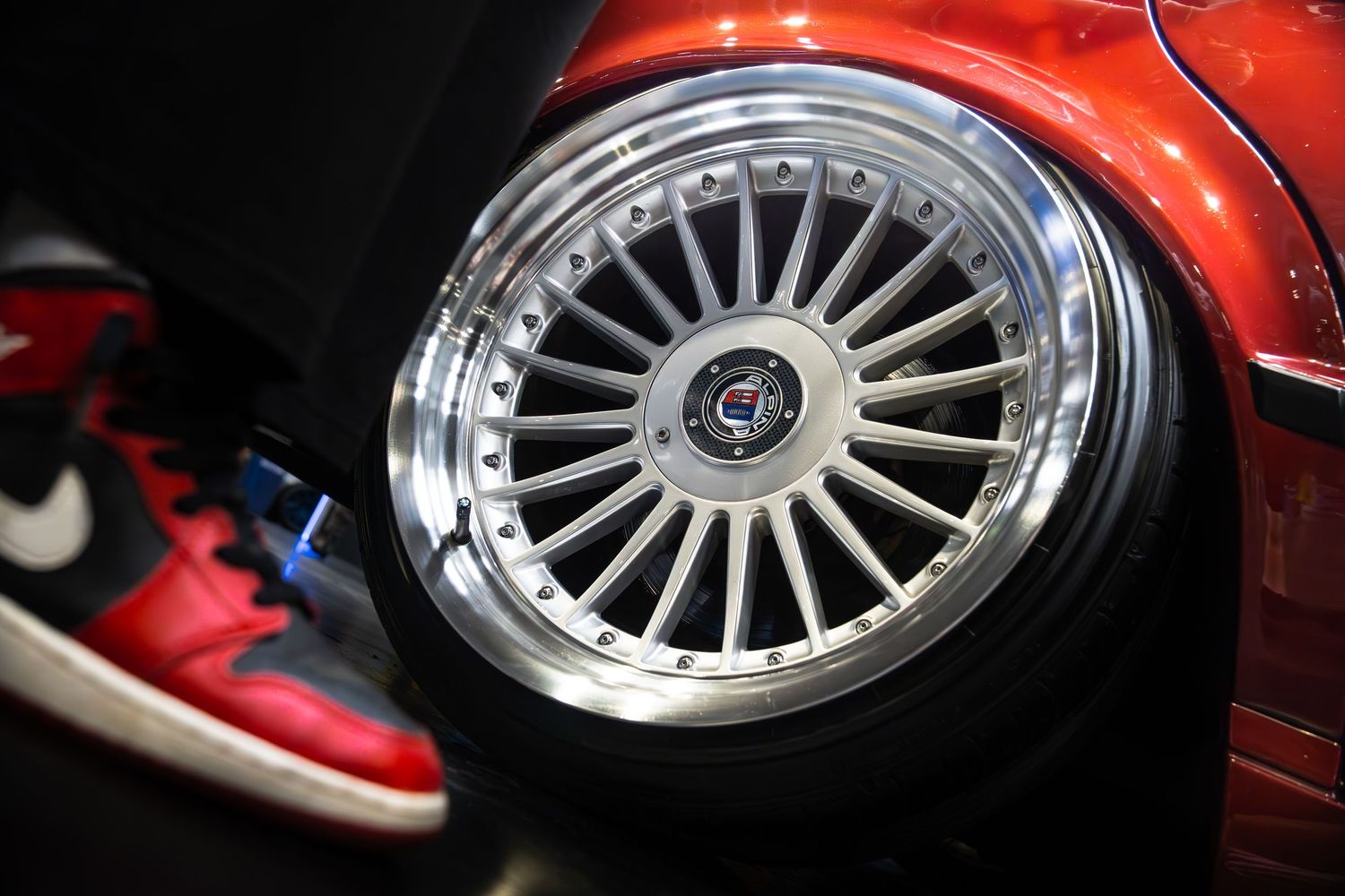 Close-up of a polished multi-spoke alloy wheel on a red car beside red Nike Air Jordan sneakers.