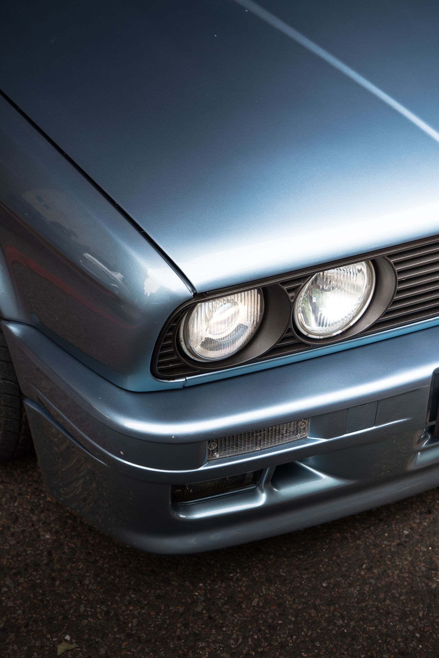 Close-up of dual round headlights on a silver-blue classic BMW E30 front bumper.