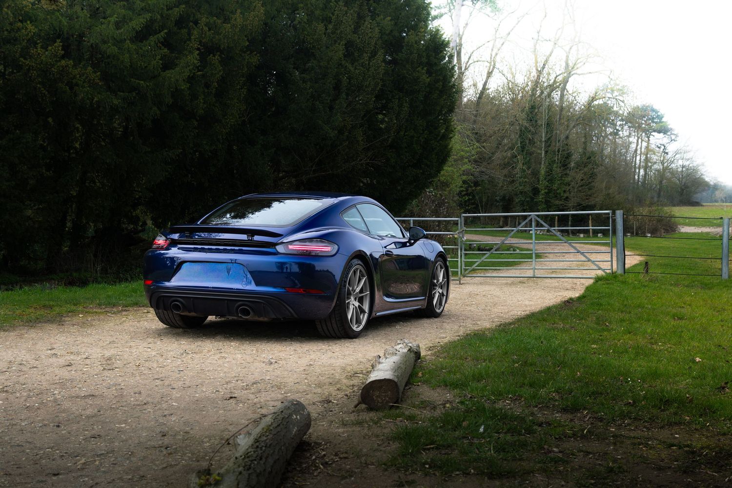Blue Porsche 718 Cayman on a gravel driveway beside a metal gate with trees in the background.