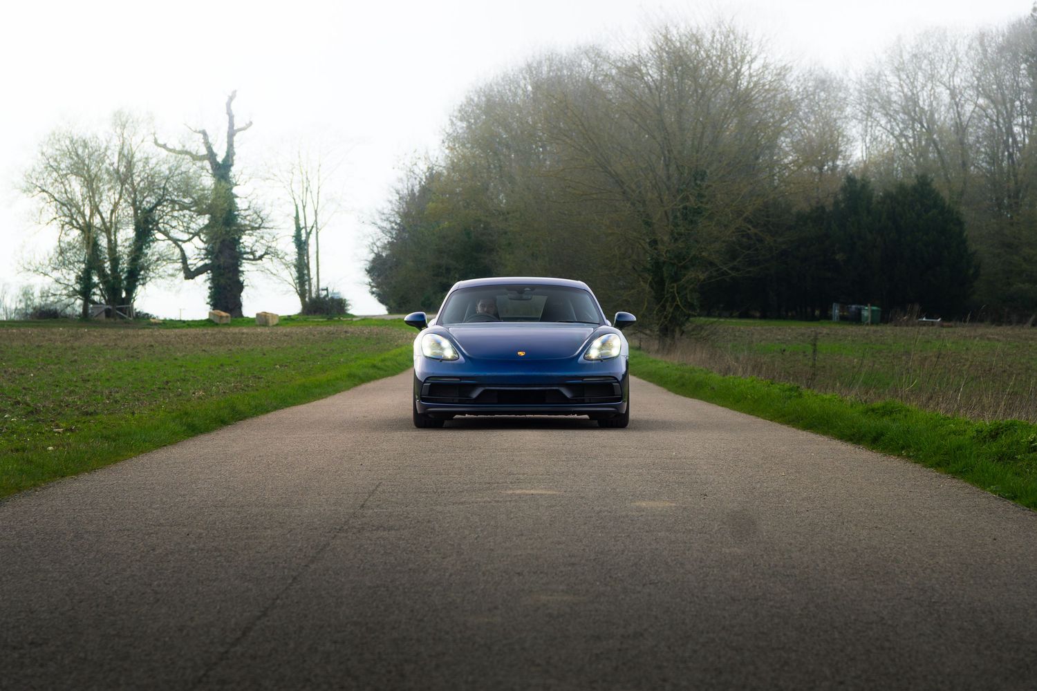 Blue Porsche 718 Cayman photographed head-on down a tree-lined country lane.