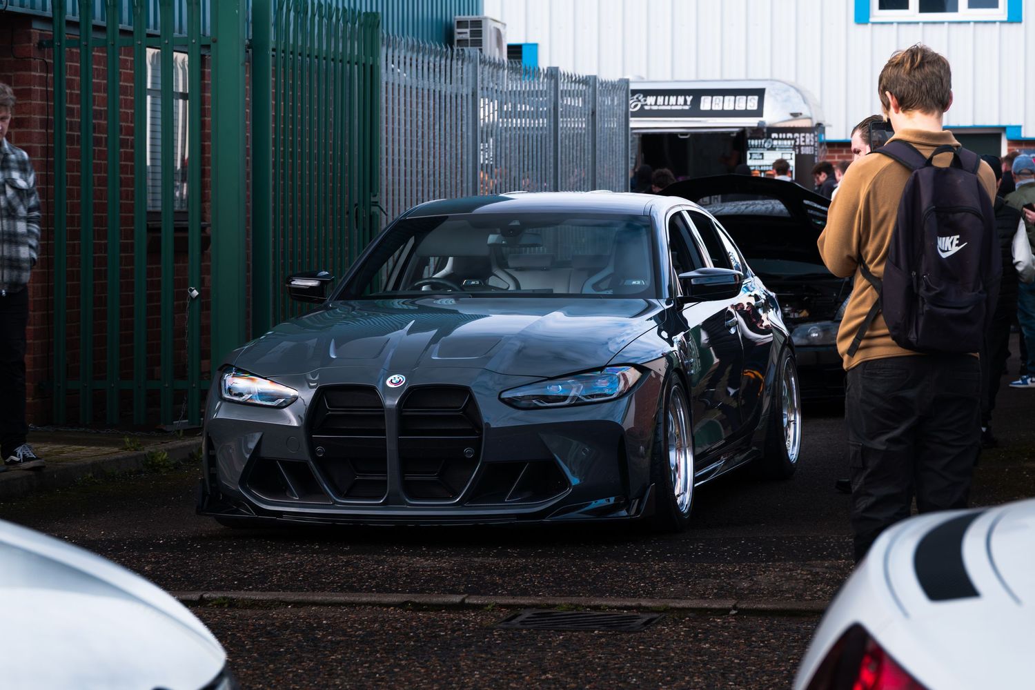 Dark grey BMW M3 G80 with wide body kit at a car meet in an industrial area.