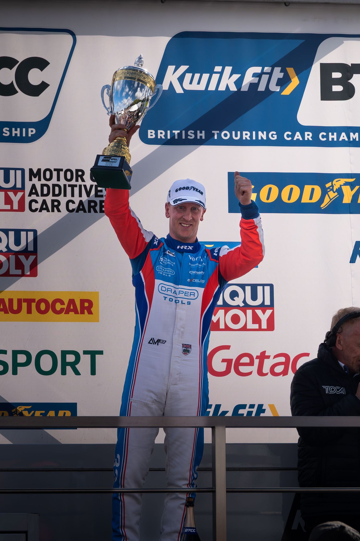 A racing driver in red and blue livery raises a trophy on the KwikFit BTCC podium in celebration.