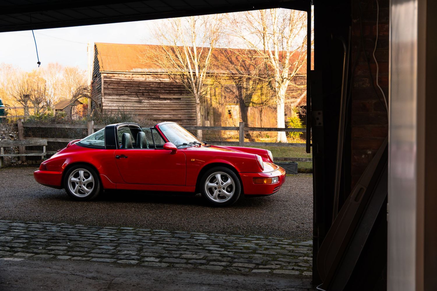 Classic red Porsche 911 Targa parked on cobblestones outside a rustic wooden barn at sunset.