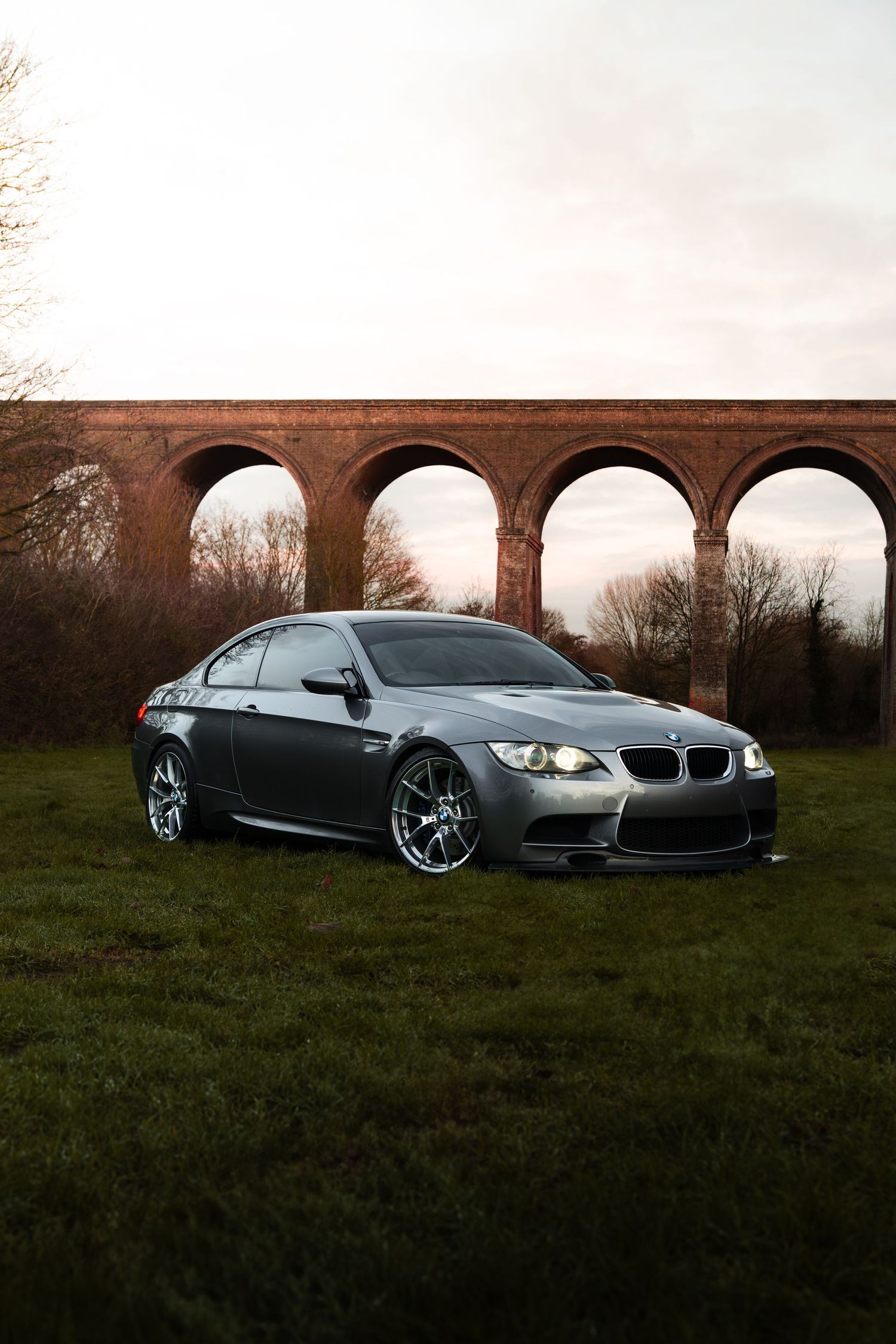 BMW M3 in gunmetal grey parked on green grass with arched brick viaduct behind.