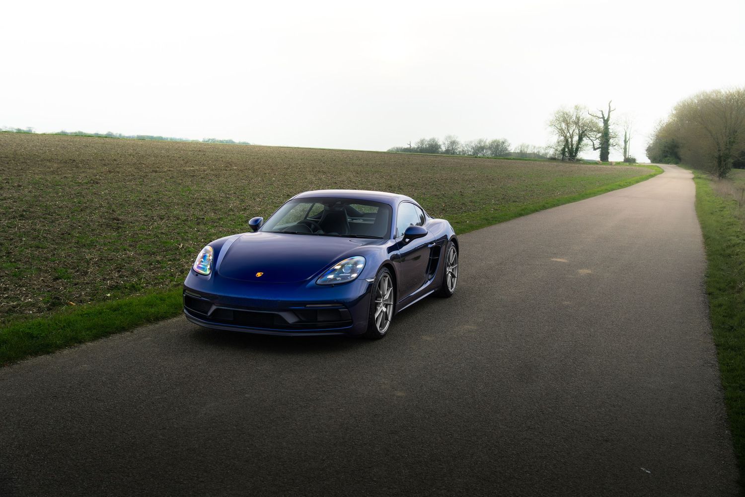 Blue Porsche 718 Cayman coupe on a rural road with green fields and overcast sky.