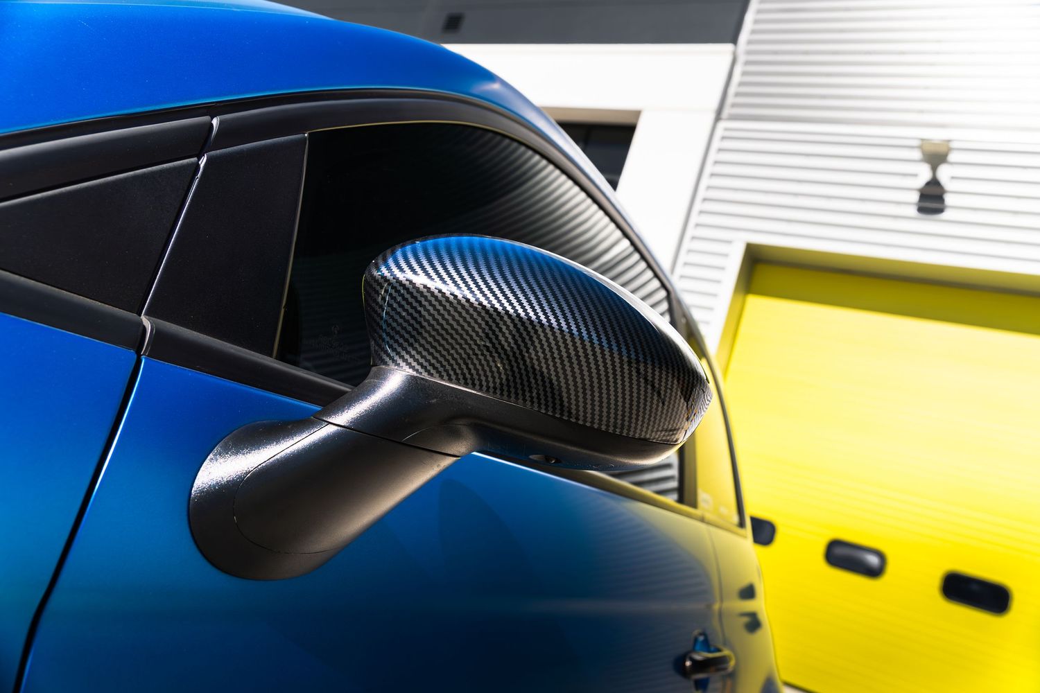 Carbon fiber wing mirror cover on a blue Abarth 500 with yellow garage in background.