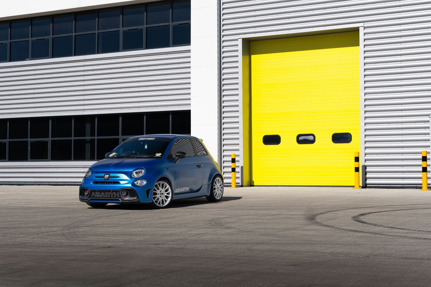 Blue Abarth 500 side profile shot in front of bright yellow industrial garage doors.