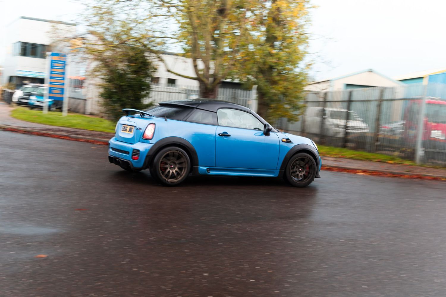 A blue Mini Cooper Coupe with bronze wheels driving on a wet urban road.