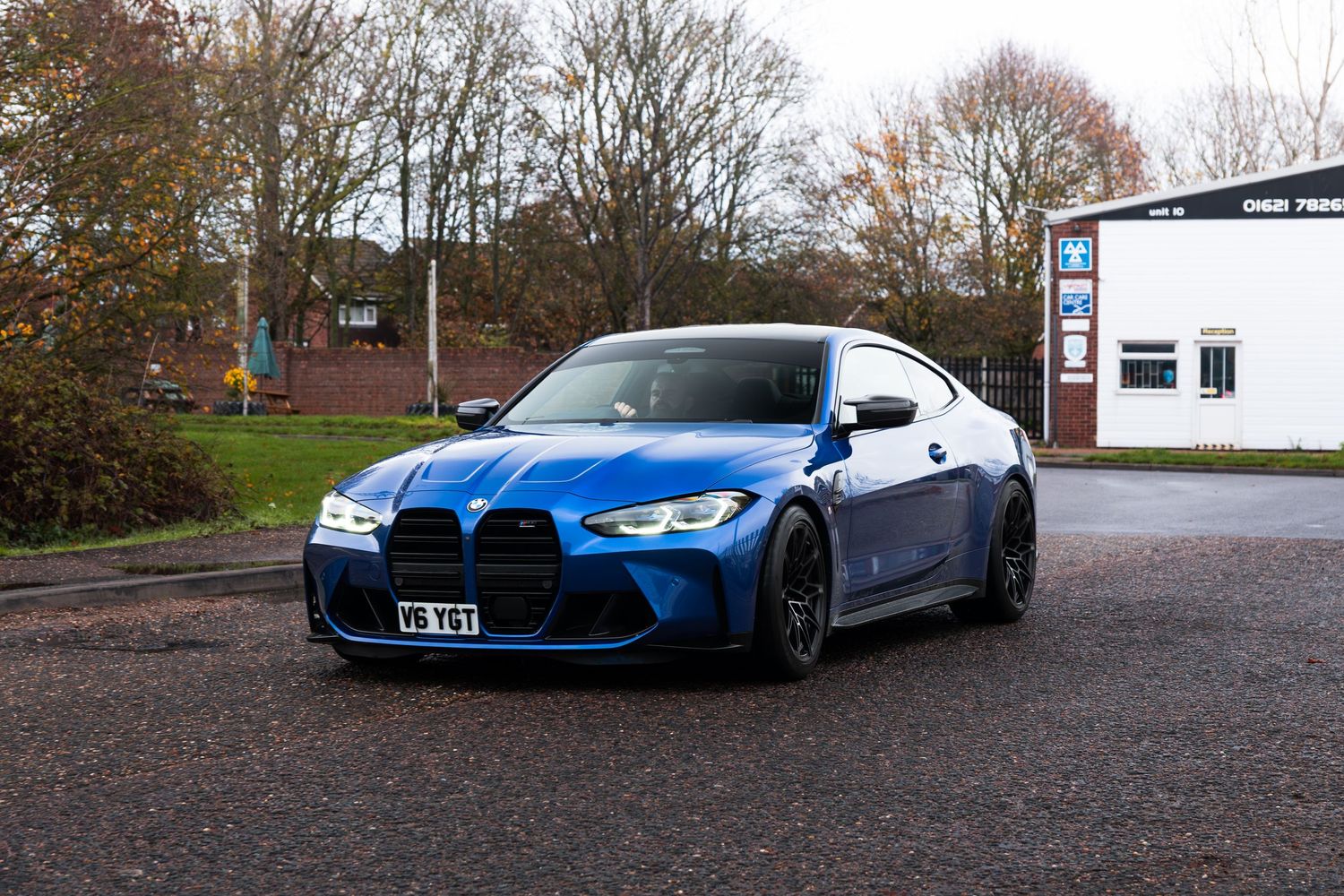 Blue BMW M4 G82 front three-quarter view parked outside UK industrial building in autumn.