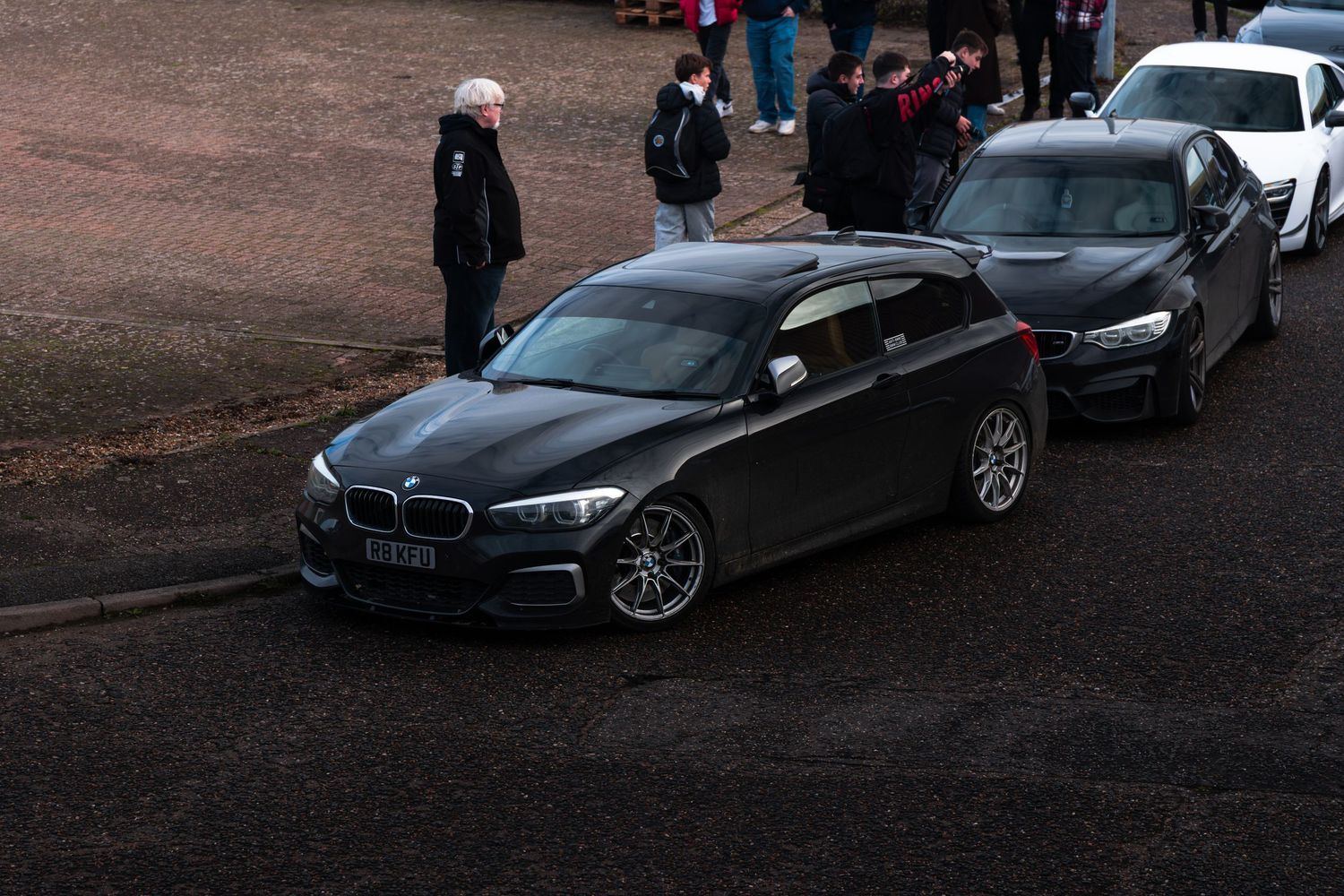 Black BMW 1 Series F20 parked on a road with spectators at a car meet event.