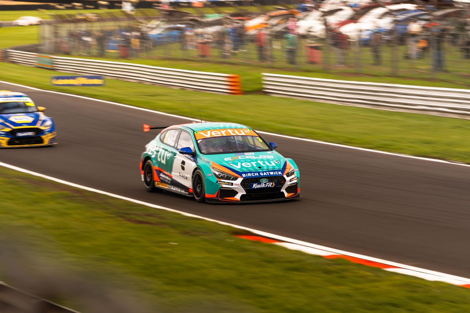 Teal and white BTCC Hyundai race car cornering at Brands Hatch circuit during race event.