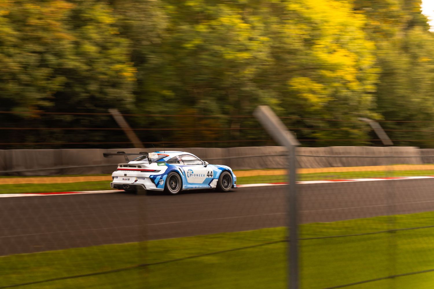 White and blue Porsche GT3 race car panning shot on track with vibrant autumn foliage backdrop.