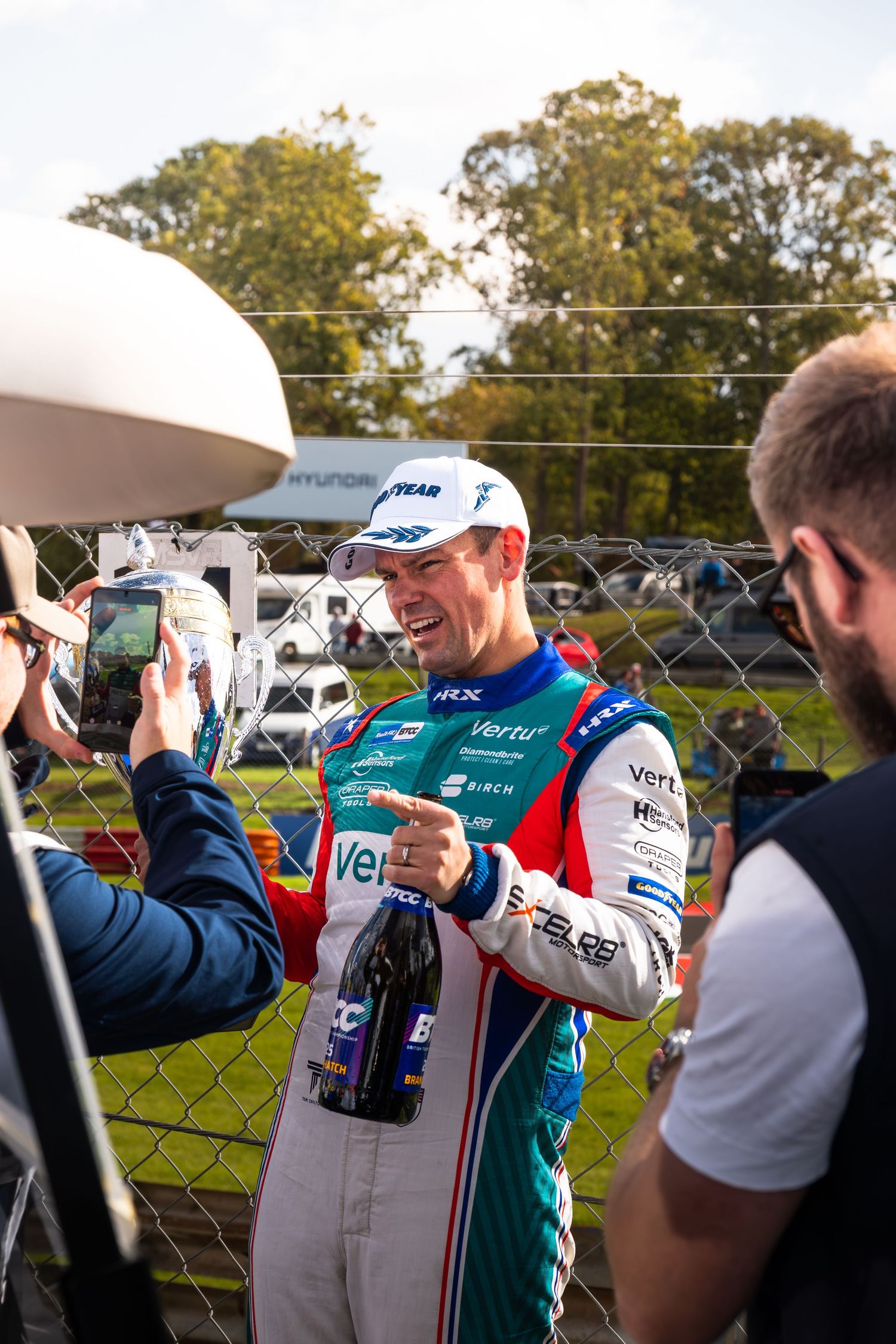A smiling BTCC racing driver in Vertu Motors livery holds a champagne bottle while speaking to media.