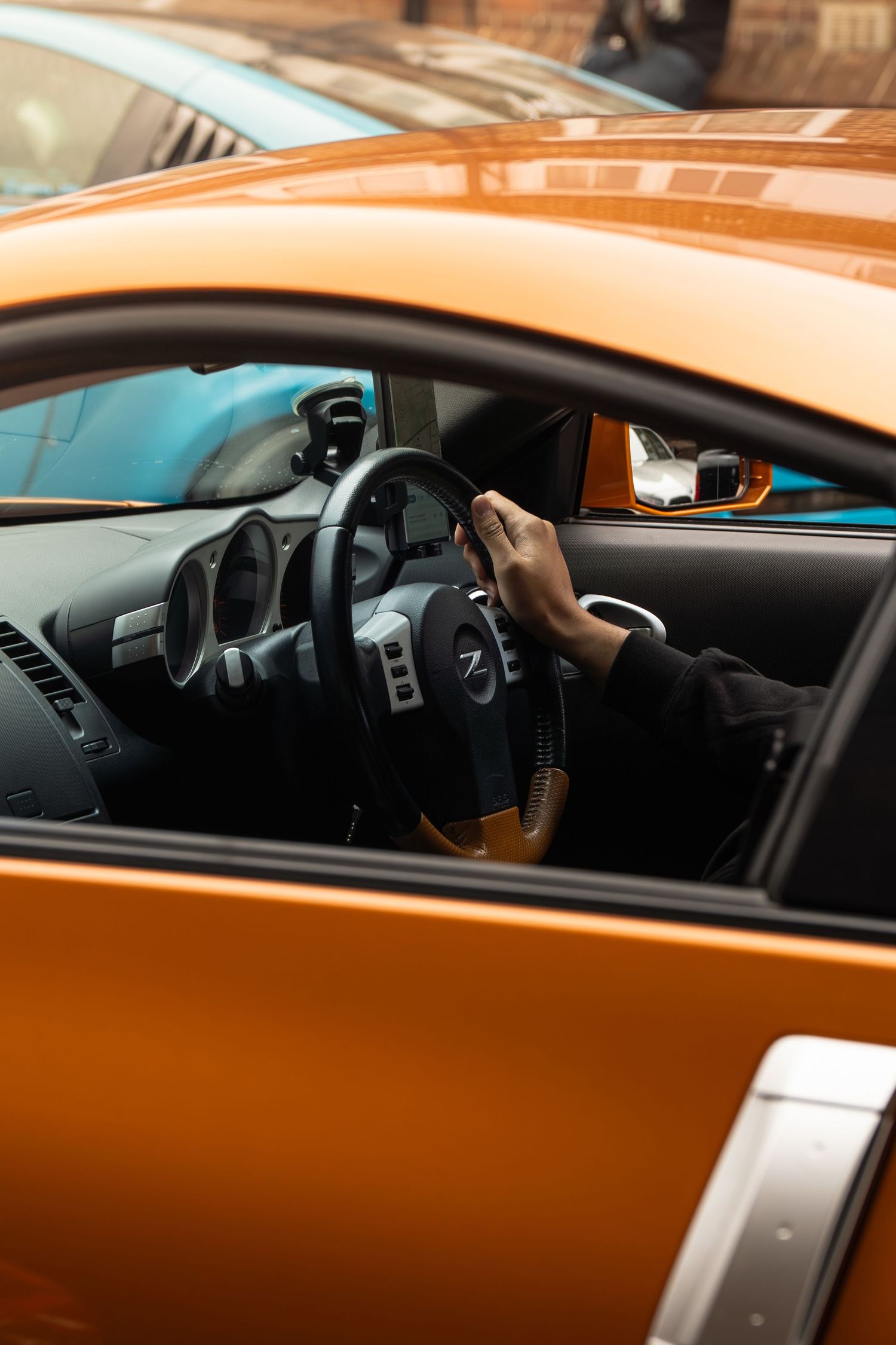 Hand gripping a Nissan steering wheel inside an orange sports car with open door.